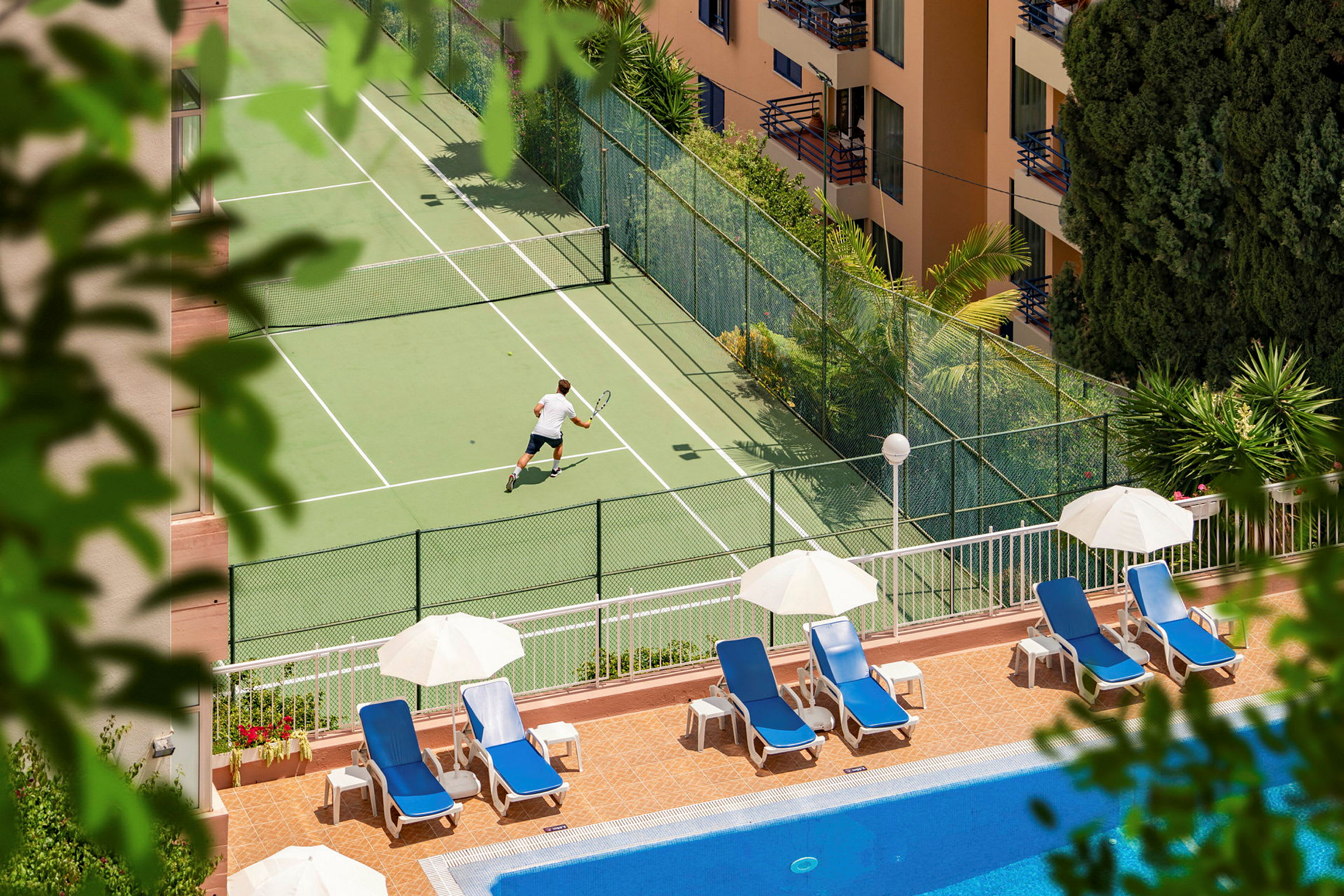 En person spiller tennis på en tennisbane nær et basseng med solsenger og parasoller. (AI-generert tekst)
