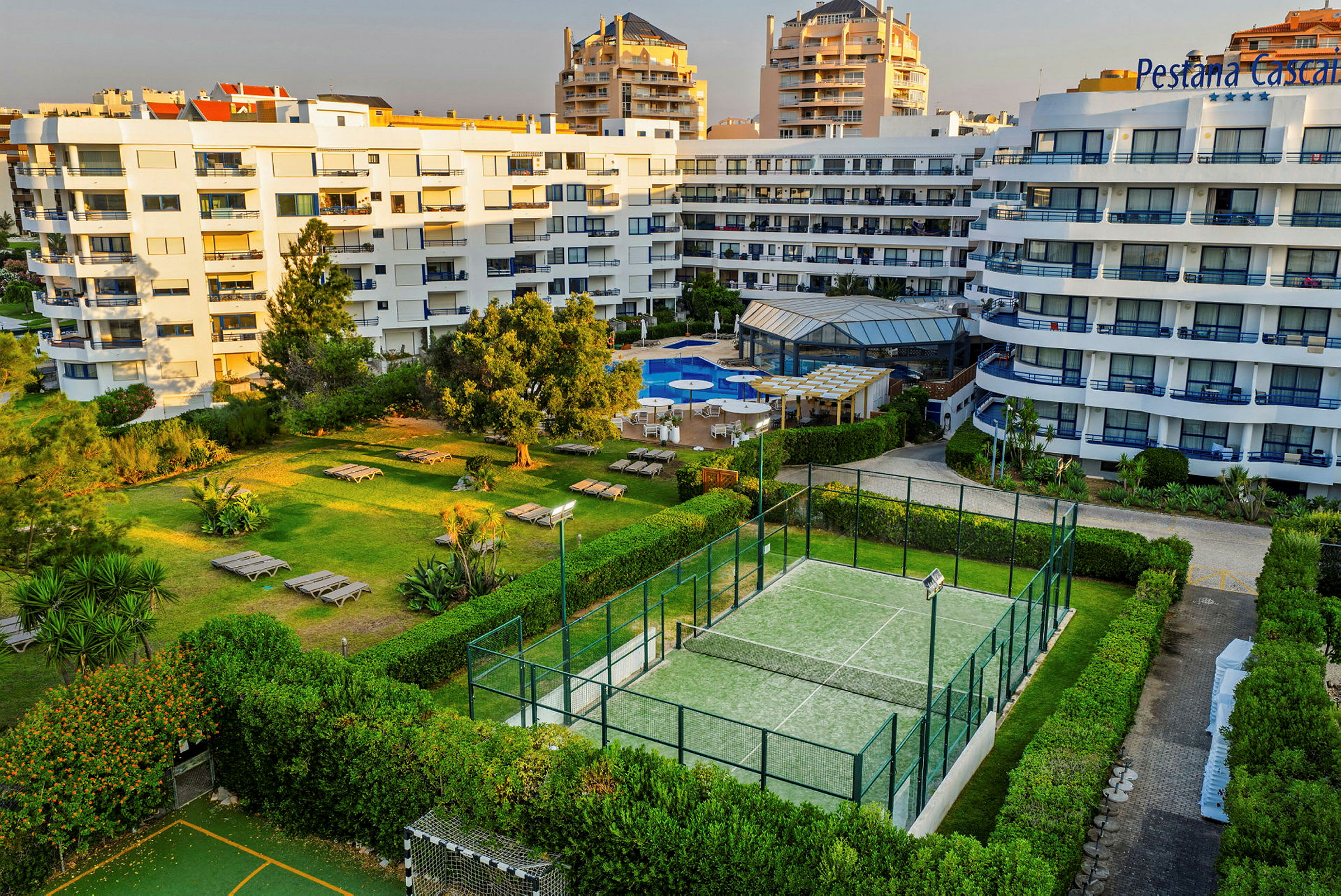 En bygning med hvit fasade, et bassengområde med solsenger og pergola samt en tennisbane omgitt av grøntområder og busker. (AI-generert tekst)