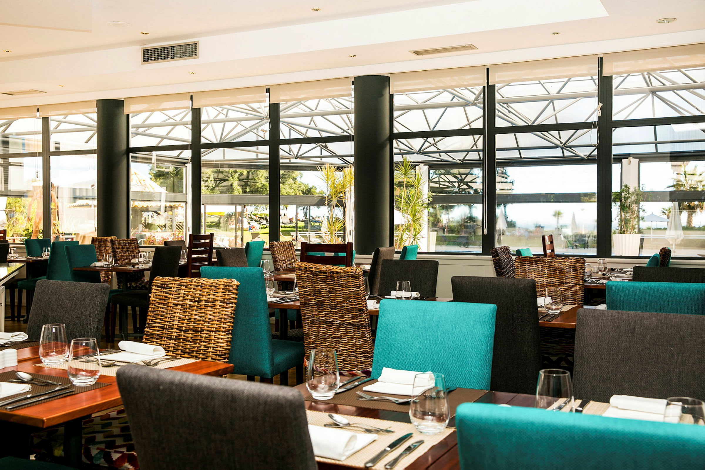 Bilde av en lys og moderne restaurant med turkise og grå stoler, dekkede bord med bestikk og tallerkener, samt store vinduer som slipper inn naturlig lys. (AI-generert tekst)