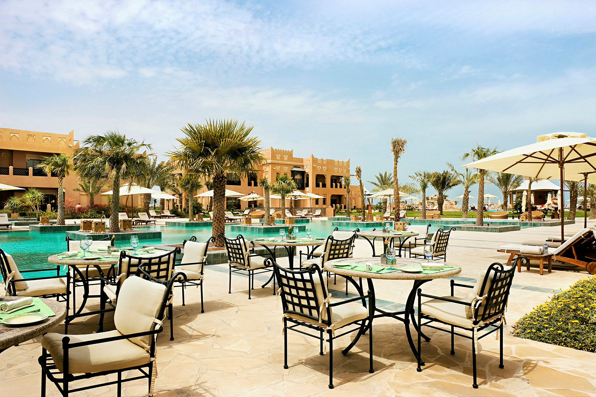 Utendørs servering ved et basseng i en hageomgivelse, omgitt av palmer og bygninger. (AI-generert tekst)