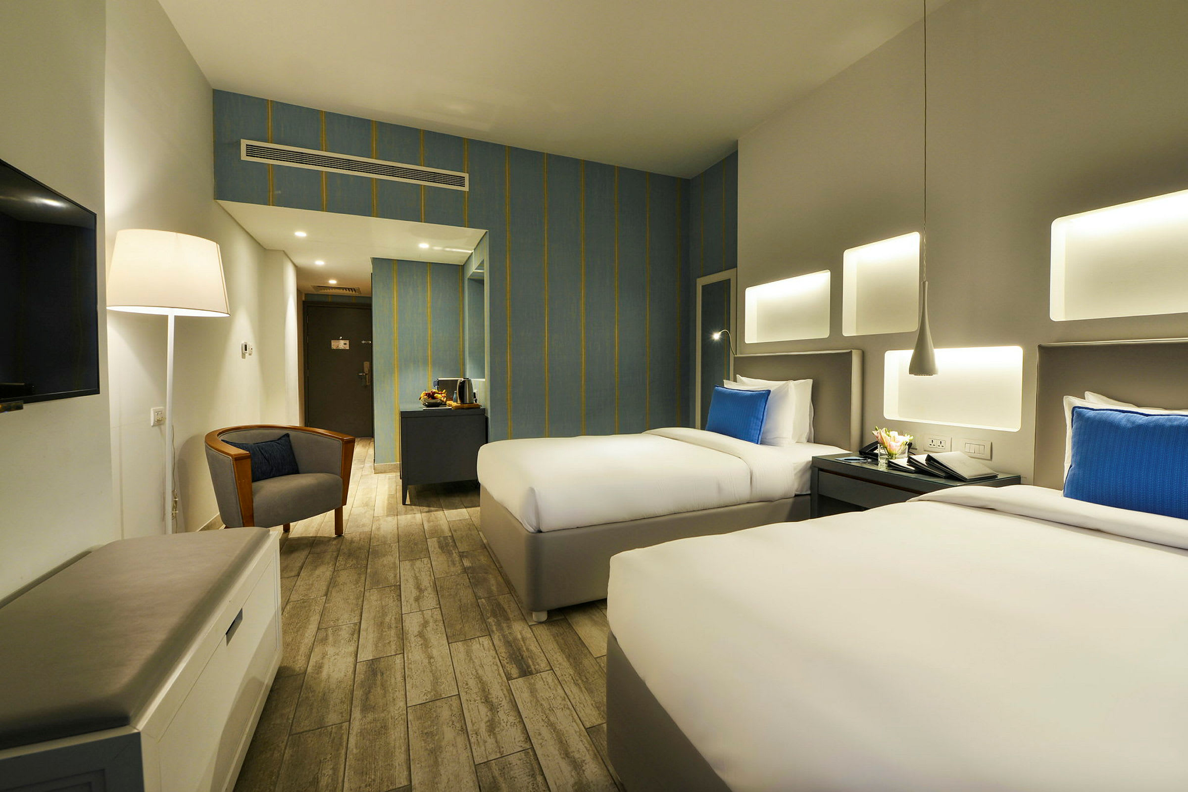 Et hotellrom med to enkeltsenger, blå puter, et skrivebord, en lesestol og en lampe. (AI-generert tekst)