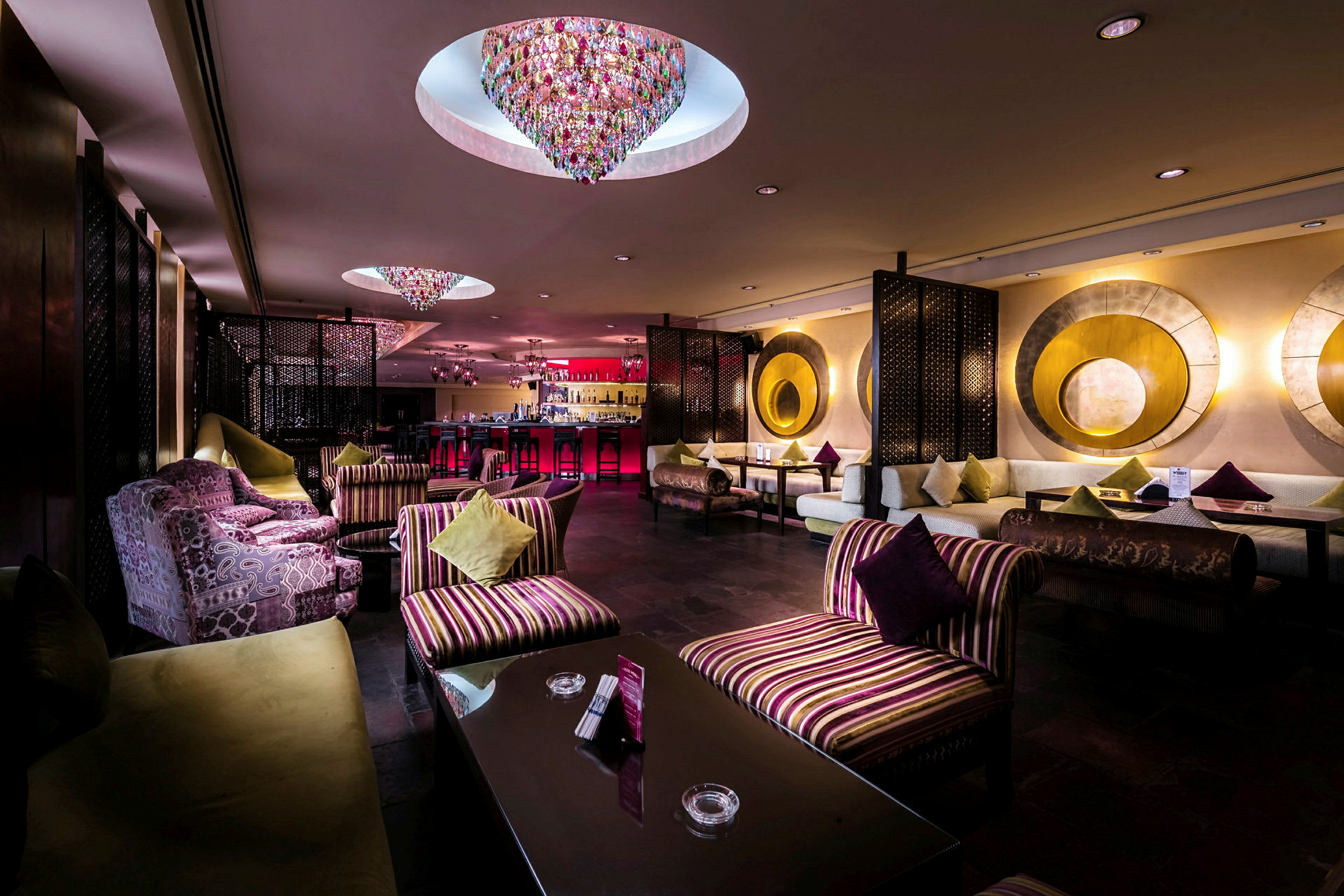 En elegant hotellbar med fargerike lysekroner i taket, myke sittemøbler og sofaer dekorert med puter. (AI-generert tekst)