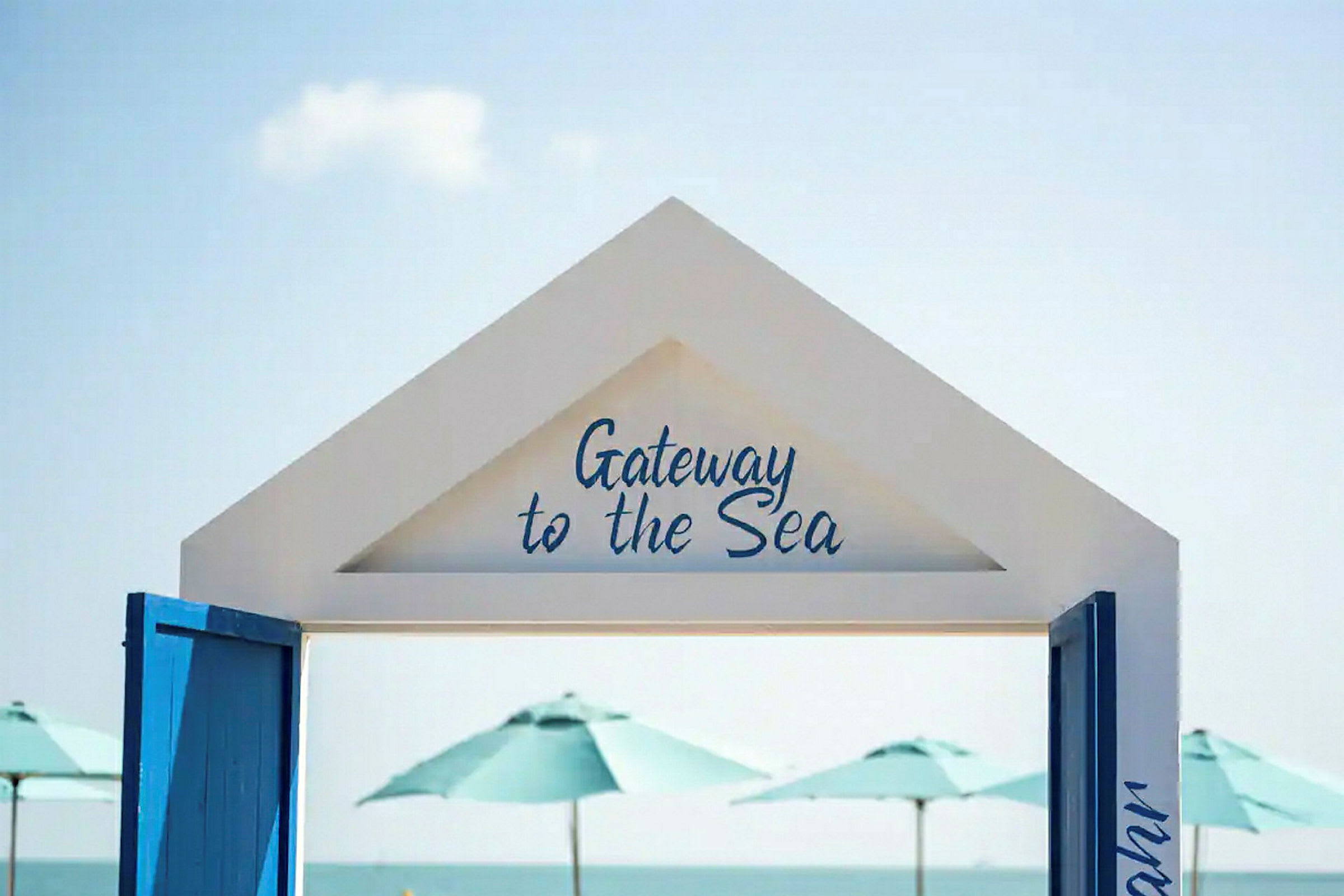 En hvit portal med teksten "Gateway to the Sea" med utsikt over havet og parasoller i bakgrunnen. (AI-generert tekst)