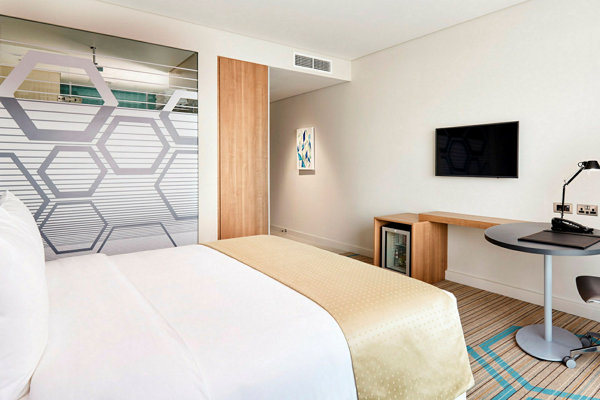 Et hotellrom med en seng dekket i hvite og beige farger. Rommet inkluderer også et skrivebord, en TV på veggen og et minikjøleskap. (AI-generert tekst)