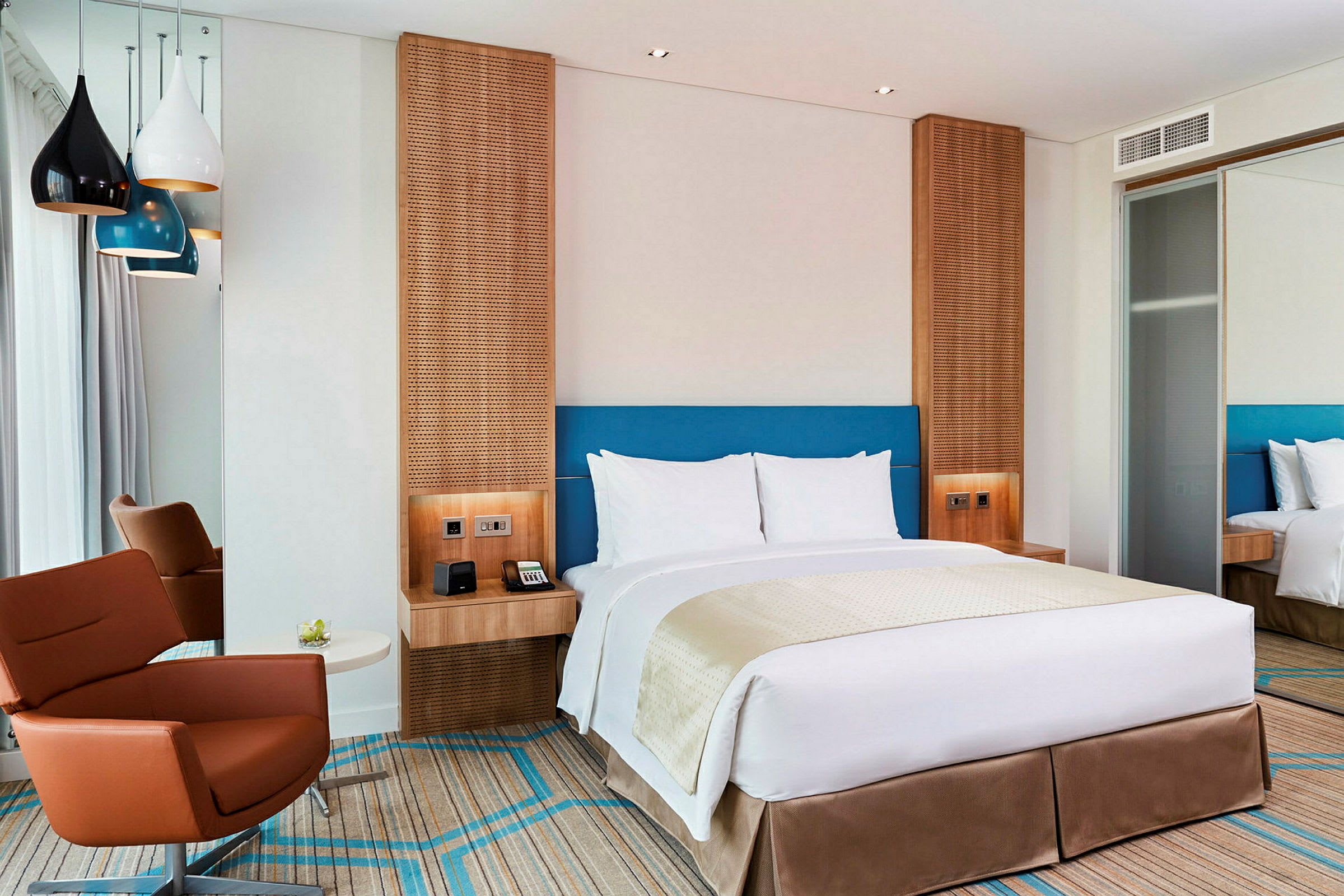 Et moderne hotellrom med en dobbeltseng, en brun lenestol og fargerike lamper som henger fra taket. (AI-generert tekst)