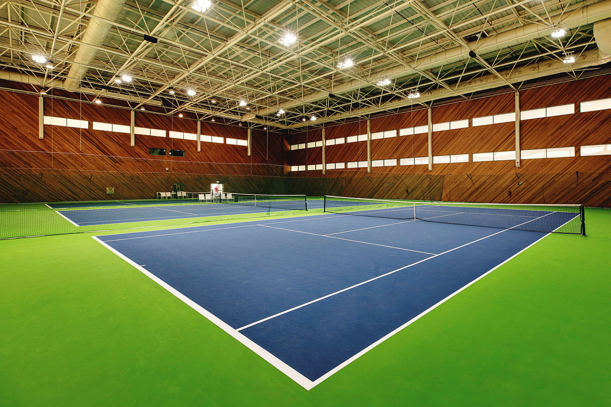 Et innendørs tennissenter med to tennisbaner, malt i blått og grønt, omgitt av trepanel på veggene. (AI-generert tekst)