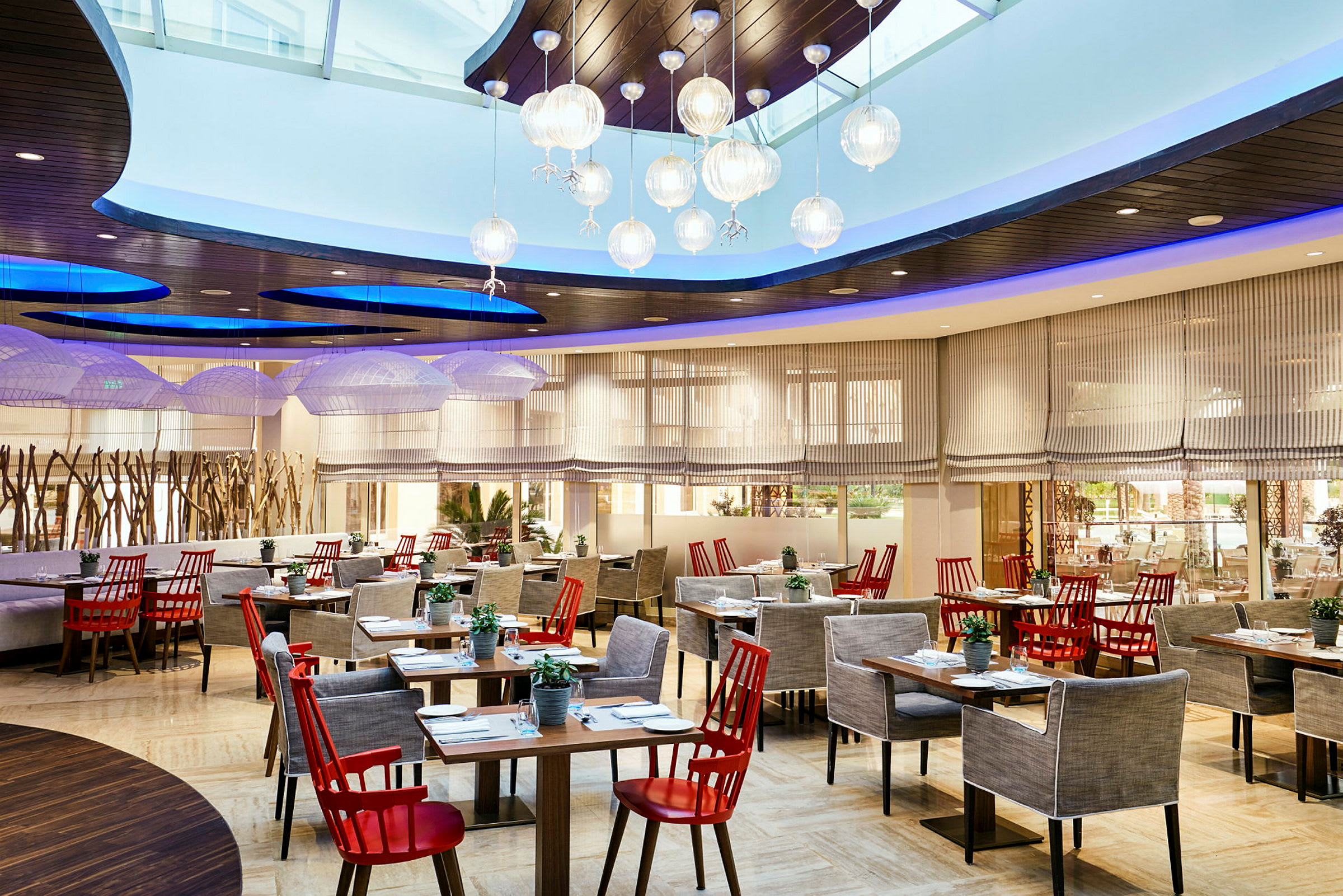 Restaurant med elegant interiør, røde stoler og grå lenestoler ved flere bord, med moderne lamper i taket og store vinduer. (AI-generert tekst)