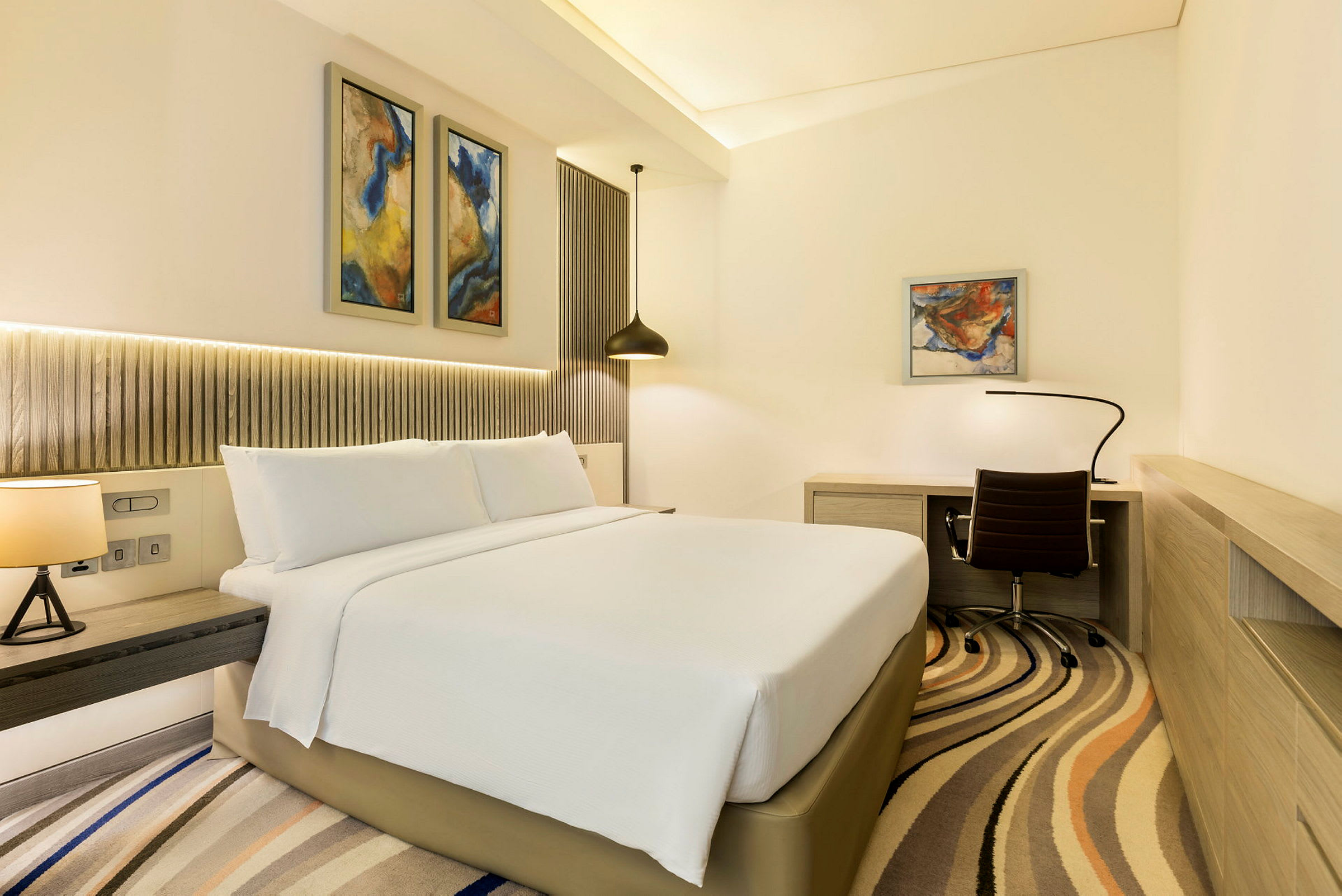 Et moderne hotellrom med en stor seng, skrivebord og stol samt abstrakt kunst på veggene. (AI-generert tekst)