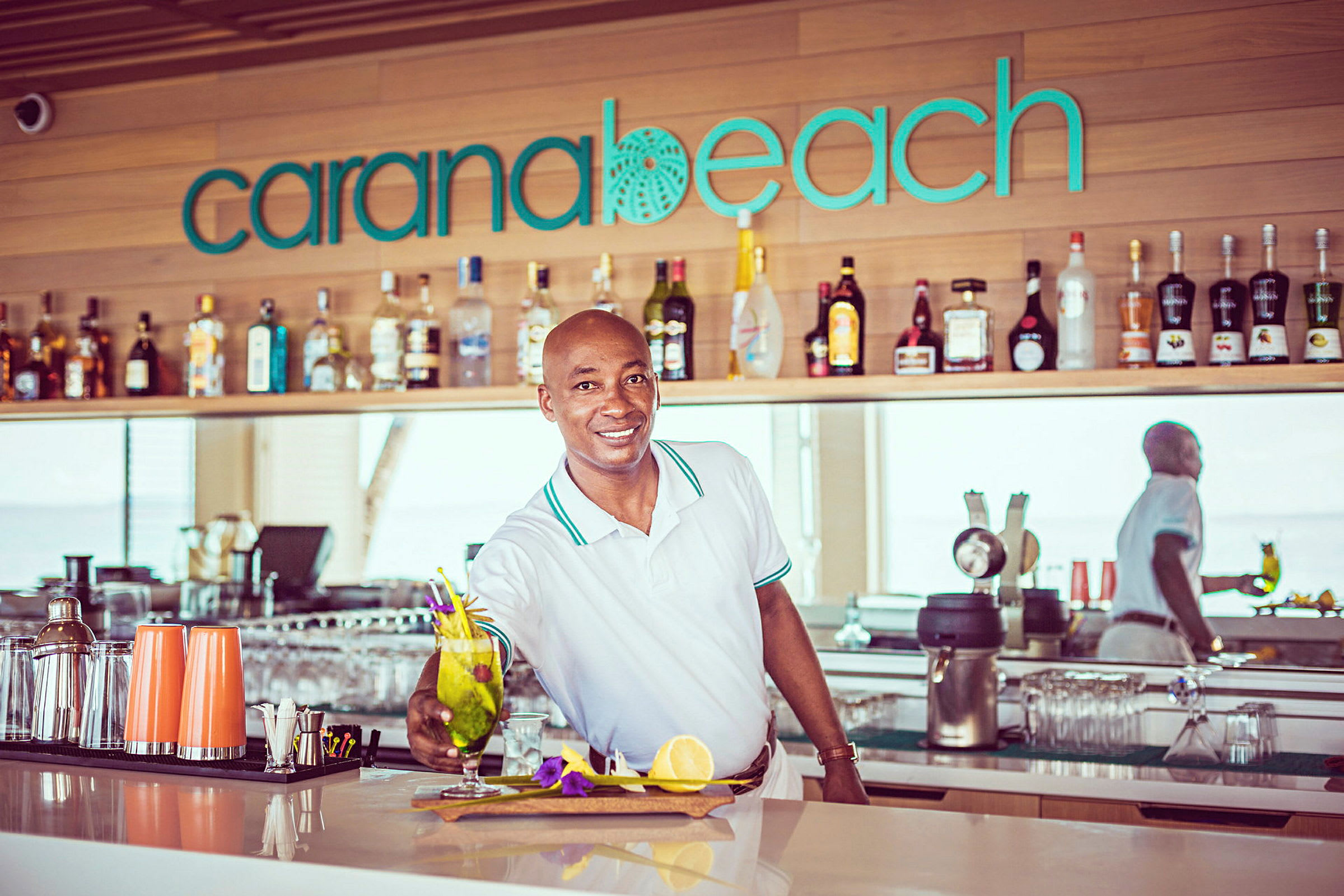 En bartender presenterer en cocktail på en bar med skiltet 'Carana Beach'. (AI-generert tekst)
