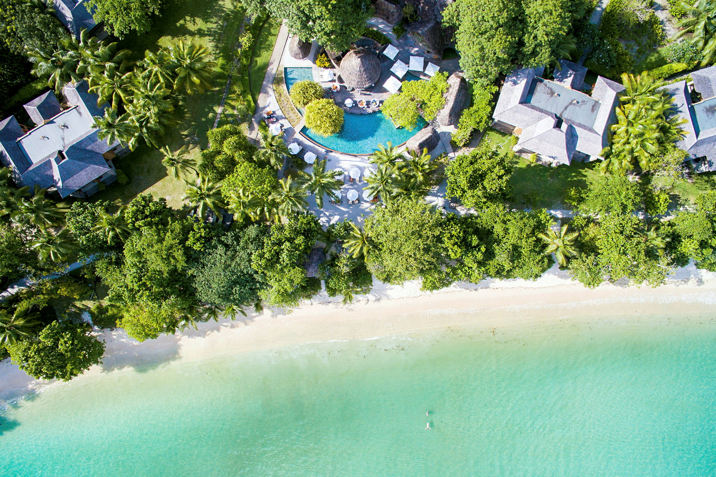 Flyfoto av en resort med basseng, bygninger omgitt av frodig vegetasjon og en strand ved turkist vann. (AI-generert tekst)