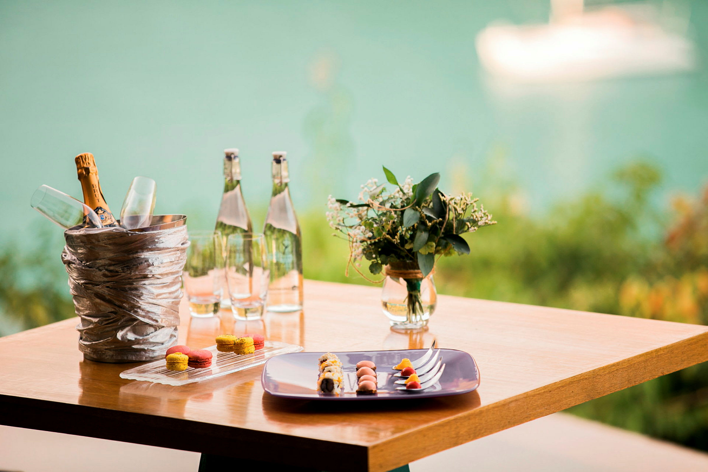 Et trebord med en champagnebøtte og glass, vannflasker, en tallerken med mat og småkaker samt en vase med blomster, med en uskarp bakgrunn av vann. (AI-generert tekst)