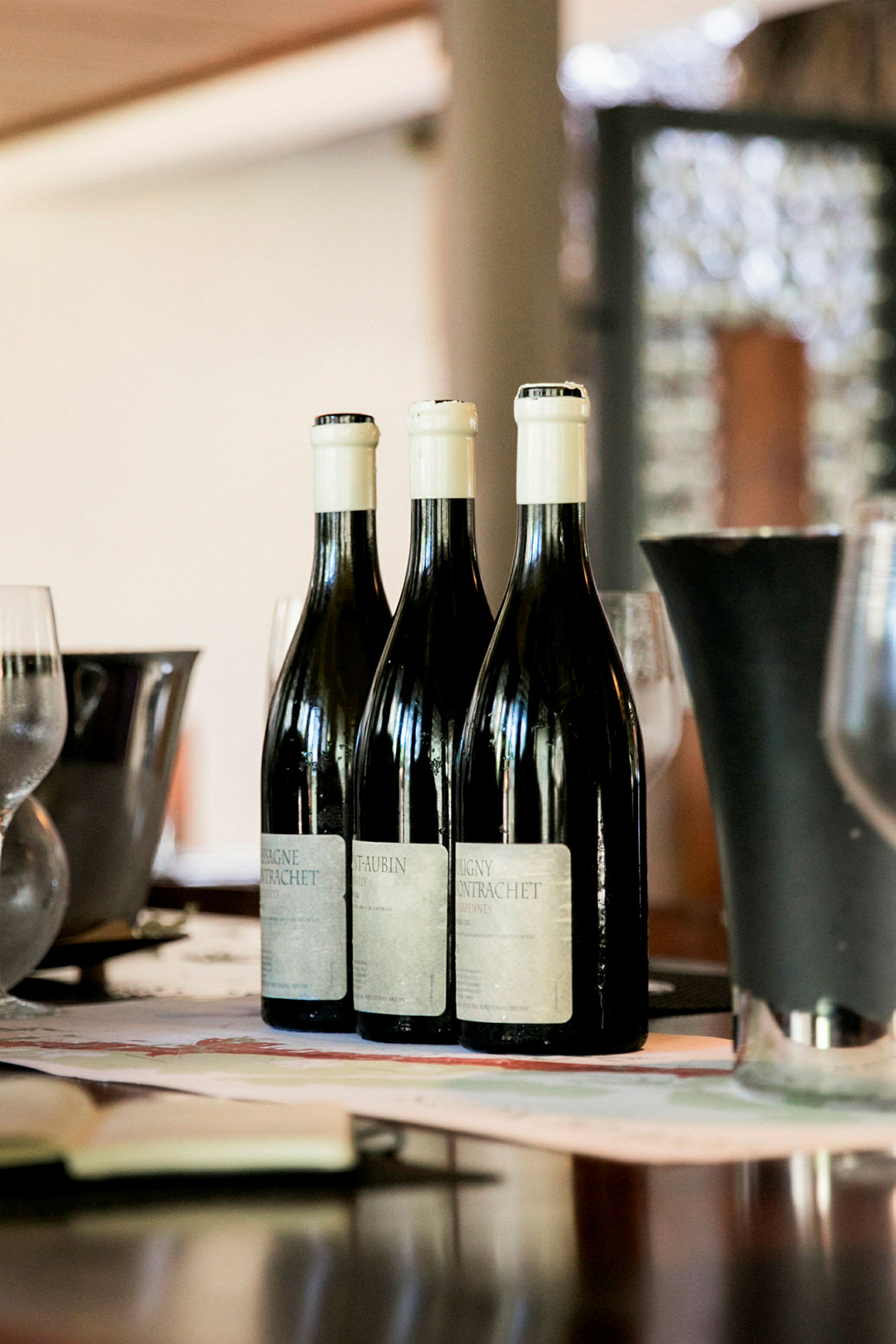 Tre vinflasker står på rekke på et bord med glass og en vase i bakgrunnen. (AI-generert tekst)