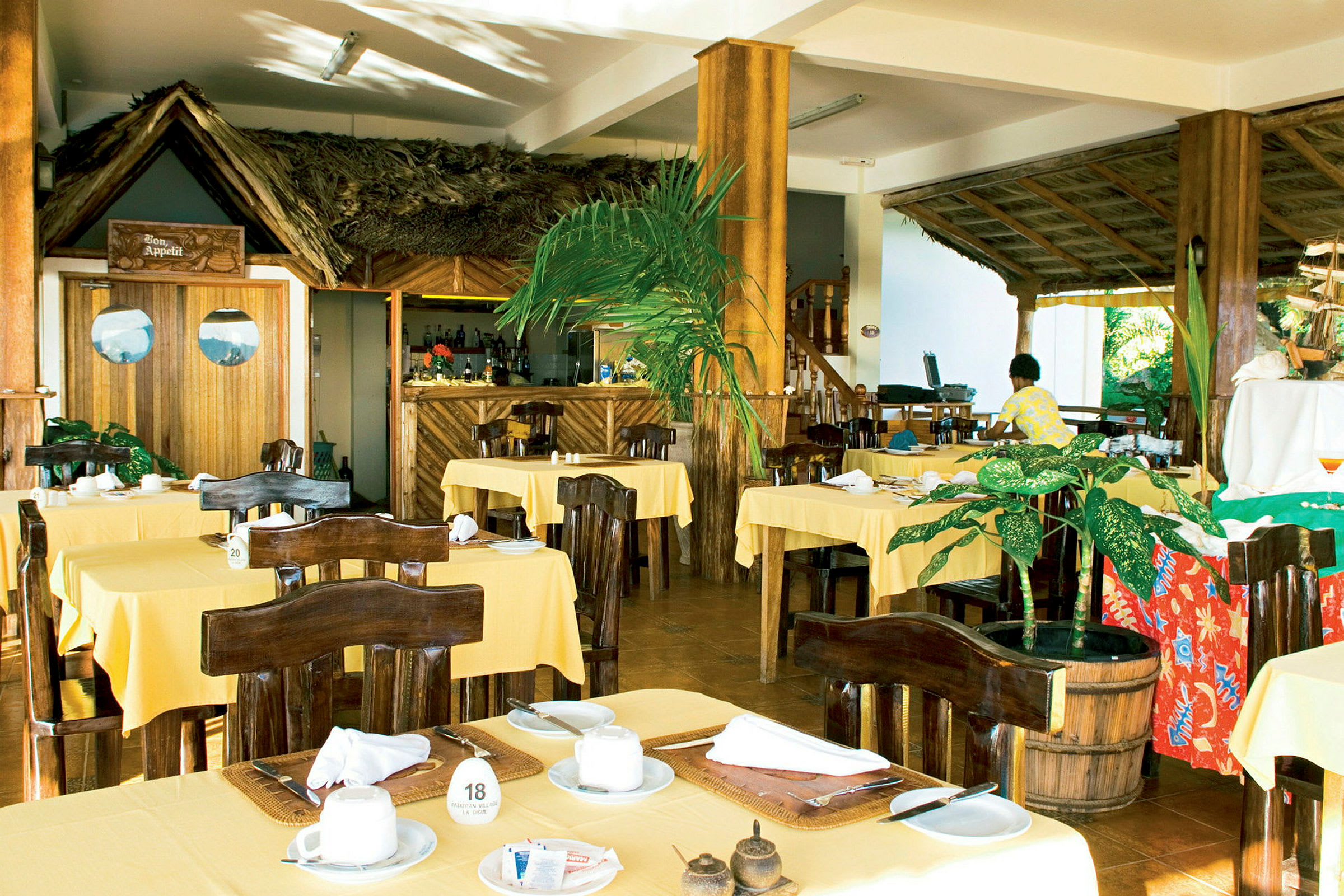 En restaurant med gule duker på bordene og mørkebrune trestoler, en bar i bakgrunnen og dekorasjoner av tropiske planter. (AI-generert tekst)