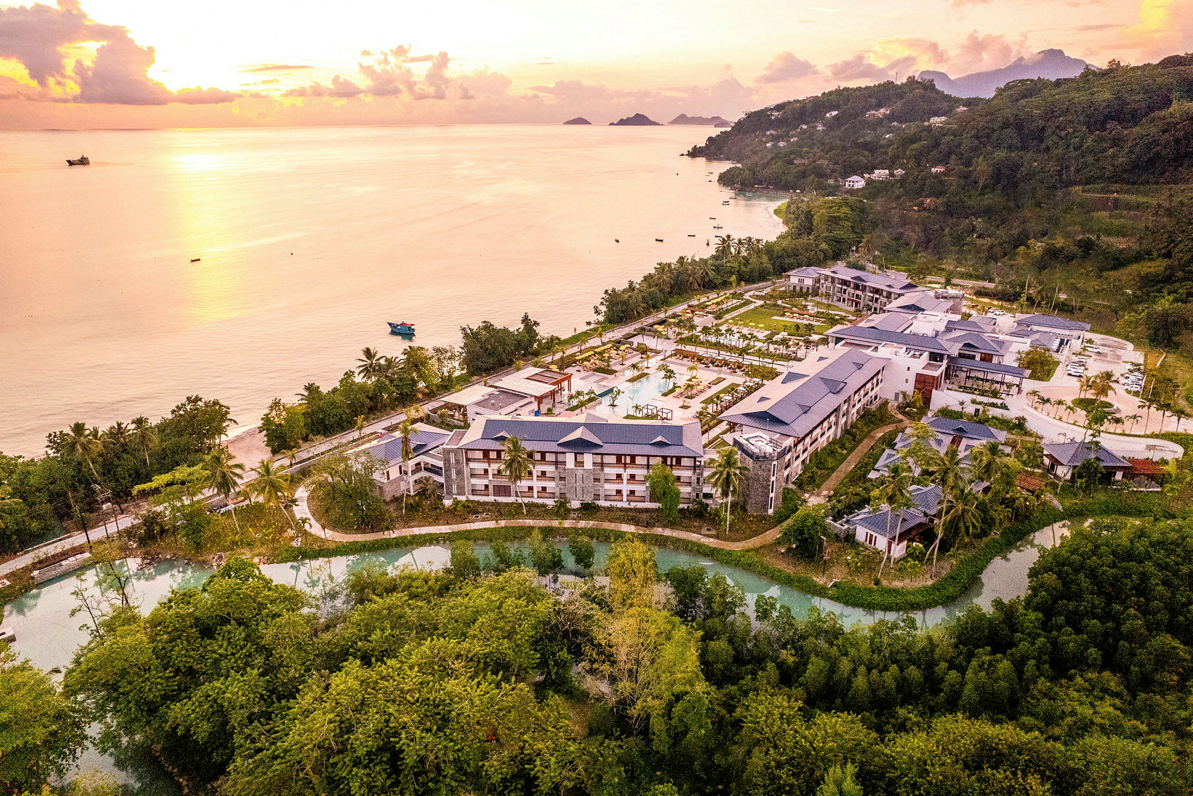 Flyfoto av en hotellkompleks omgitt av natur og plassert nær en kyst med strand og hav ved solnedgang. (AI-generert tekst)