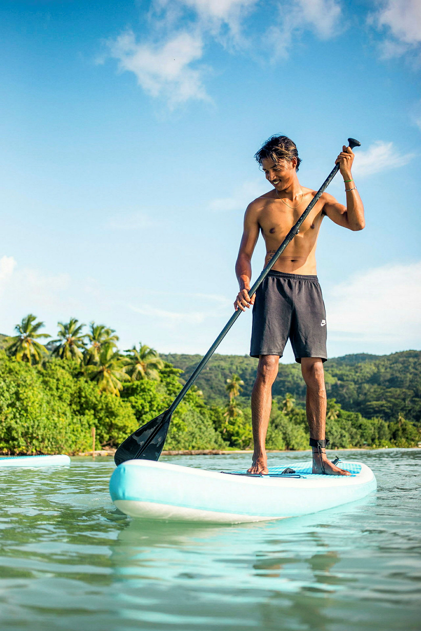 En person står på et paddleboard og padler i rolig vann med frodig vegetasjon i bakgrunnen. (AI-generert tekst)