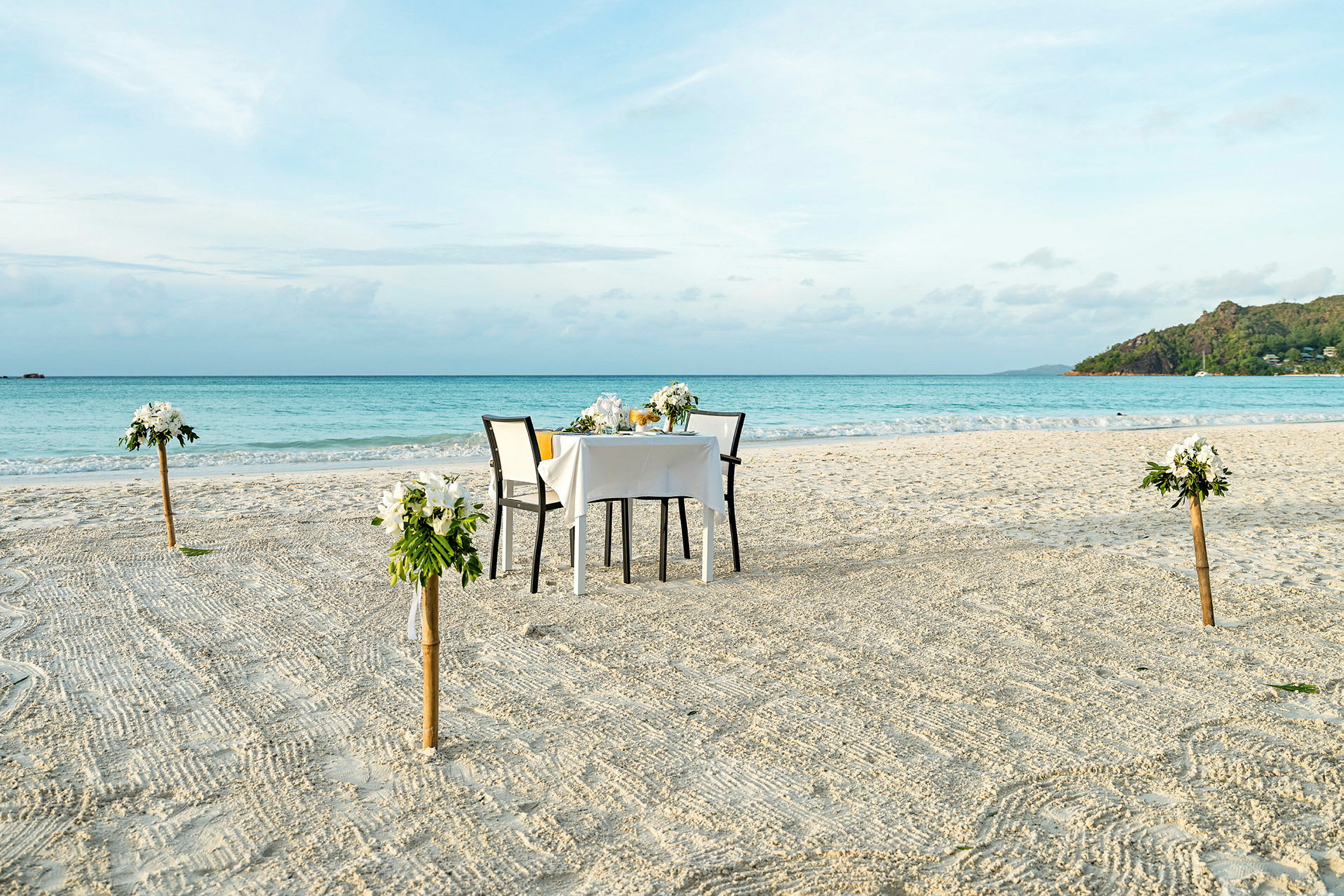 Et dekket bord med to stoler står på en sandstrand. Bordet er dekorert med blomsterdekorasjoner, og fire blomsterdekorasjoner er plassert på stolper rundt bordet. I bakgrunnen sees havet og en grønn klippe. (AI-generert tekst)