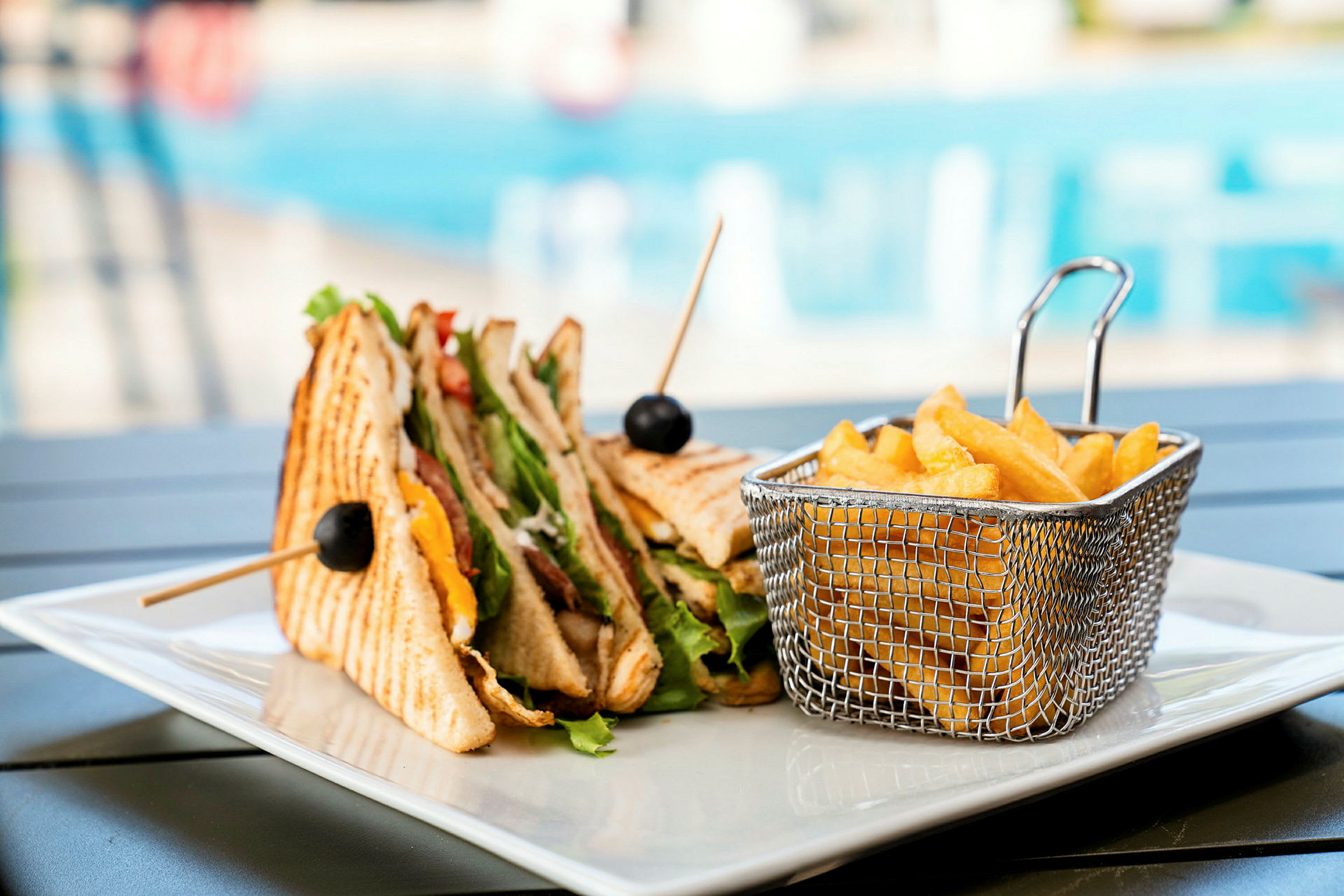 En tallerken med en trippel klubbsandwich og en kurv med pommes frites. I bakgrunnen skimtes et svømmebasseng. (AI-generert tekst)