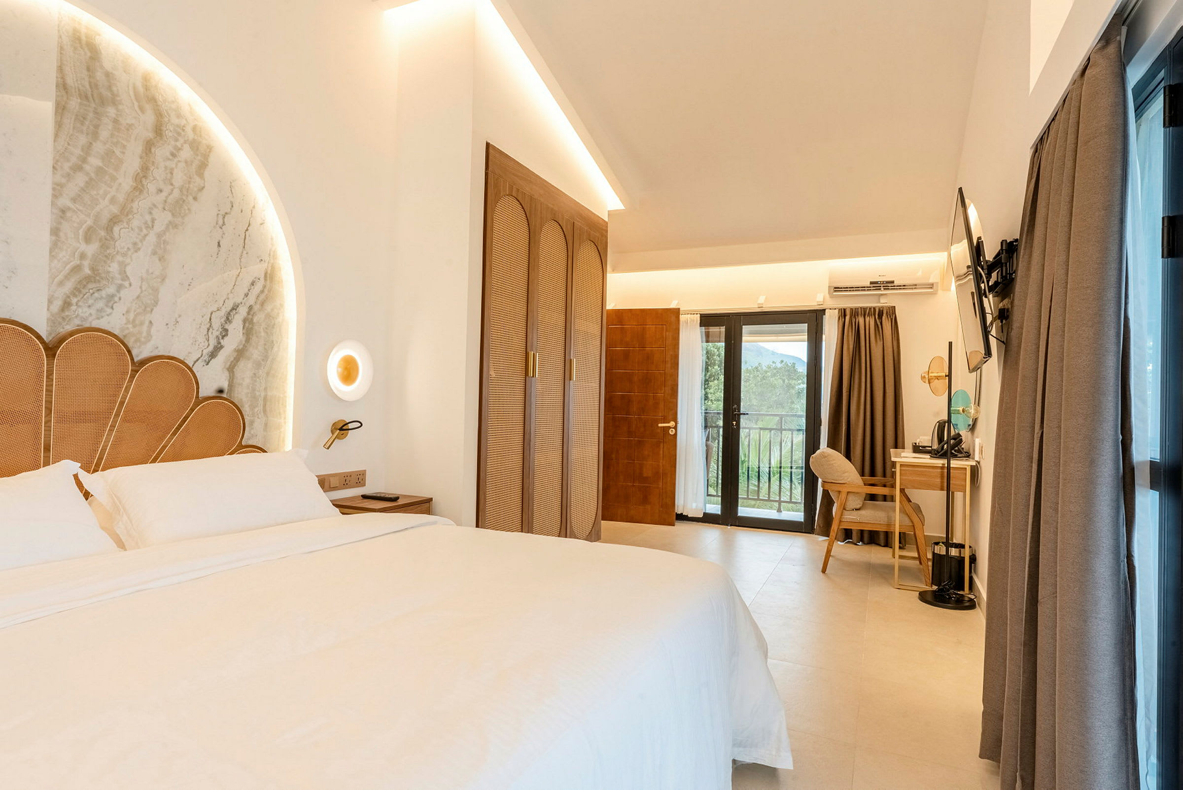 Et hotellrom med en stor seng med dekorativ sengegavl og nattbord. Garderobe, skrivebord med stol og TV, samt balkongdør med utsikt. (AI-generert tekst)