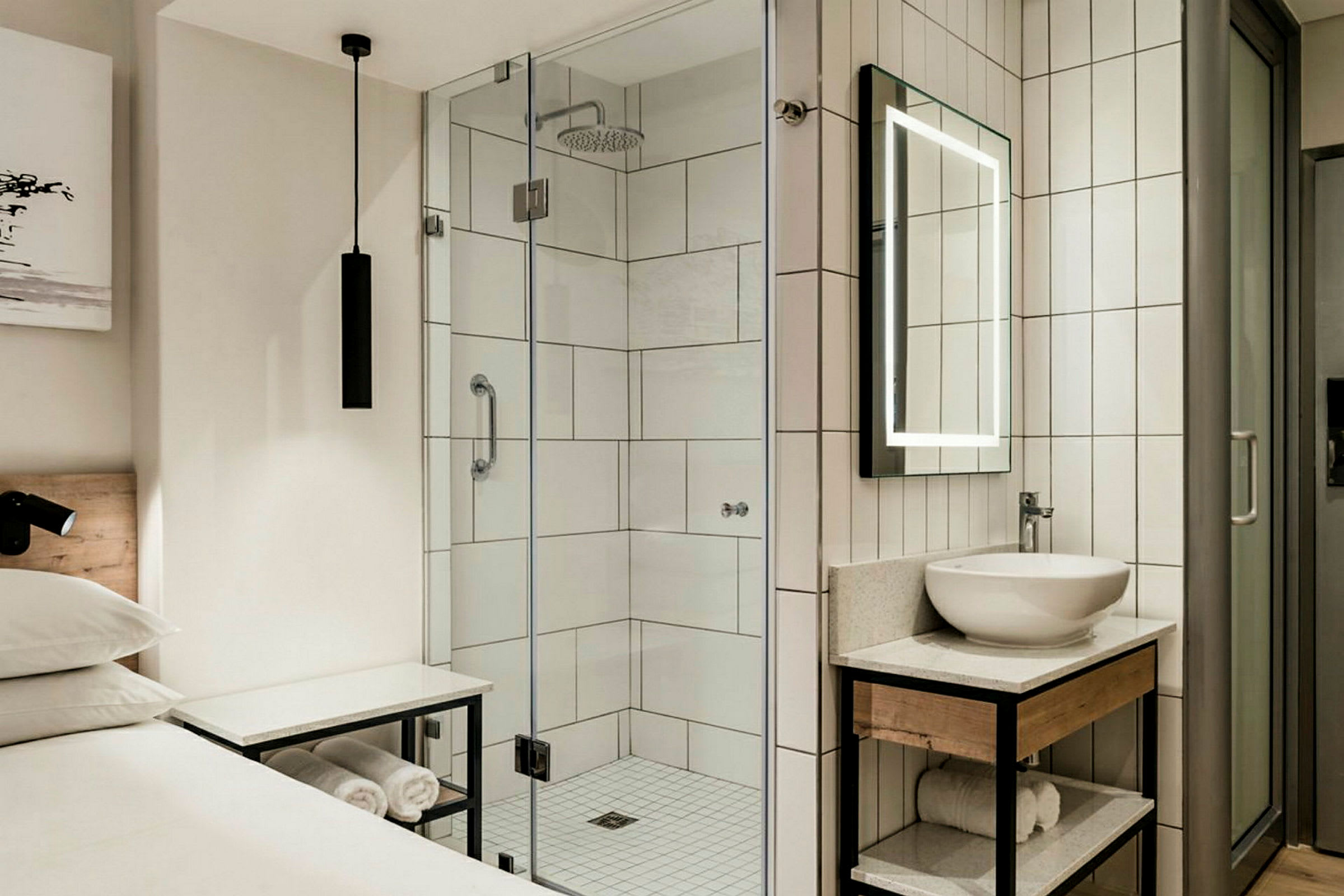 Et hotellrom med en dusj med glassvegger, et speil med belysning og en servant på en moderne møbel med håndklær under. (AI-generert tekst)