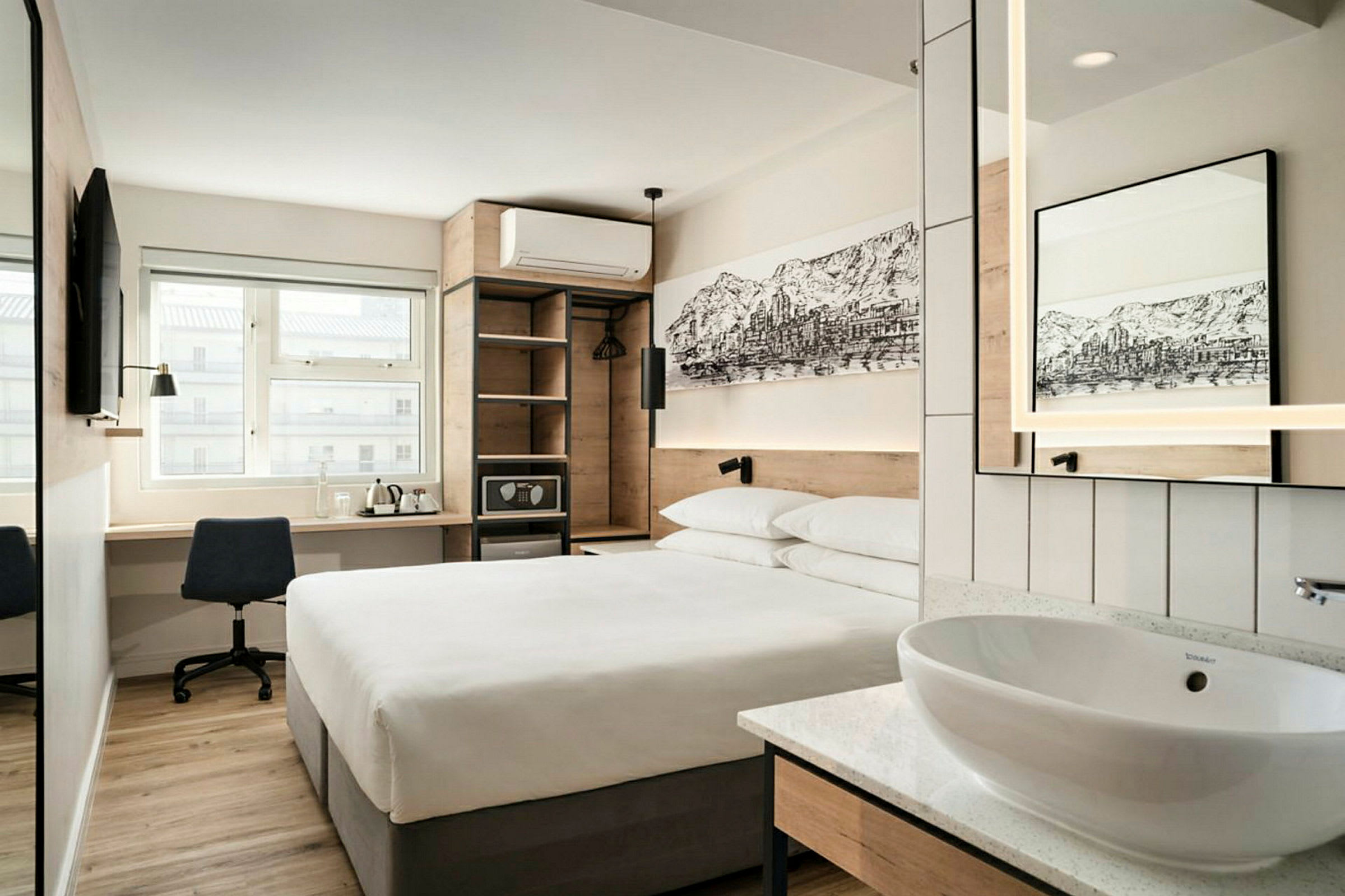 Et moderne hotellrom med en dobbeltseng, et skrivebord med stol, en veggmontert TV, samt en åpen garderobe og en liten vask med speil. (AI-generert tekst)
