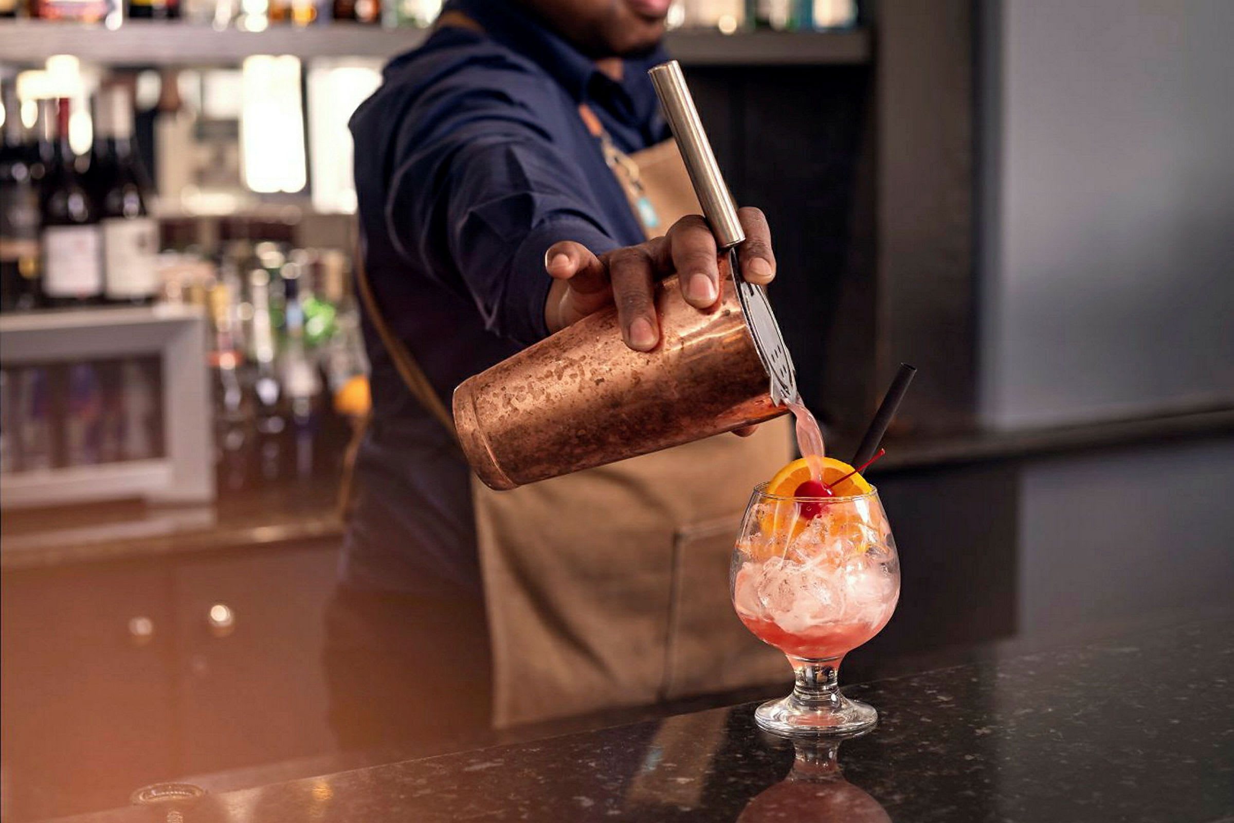 En bartender heller en rosa drikk fra en shaker i et glass med is og en dekorasjon av appelsinskive og kirsebær. (AI-generert tekst)