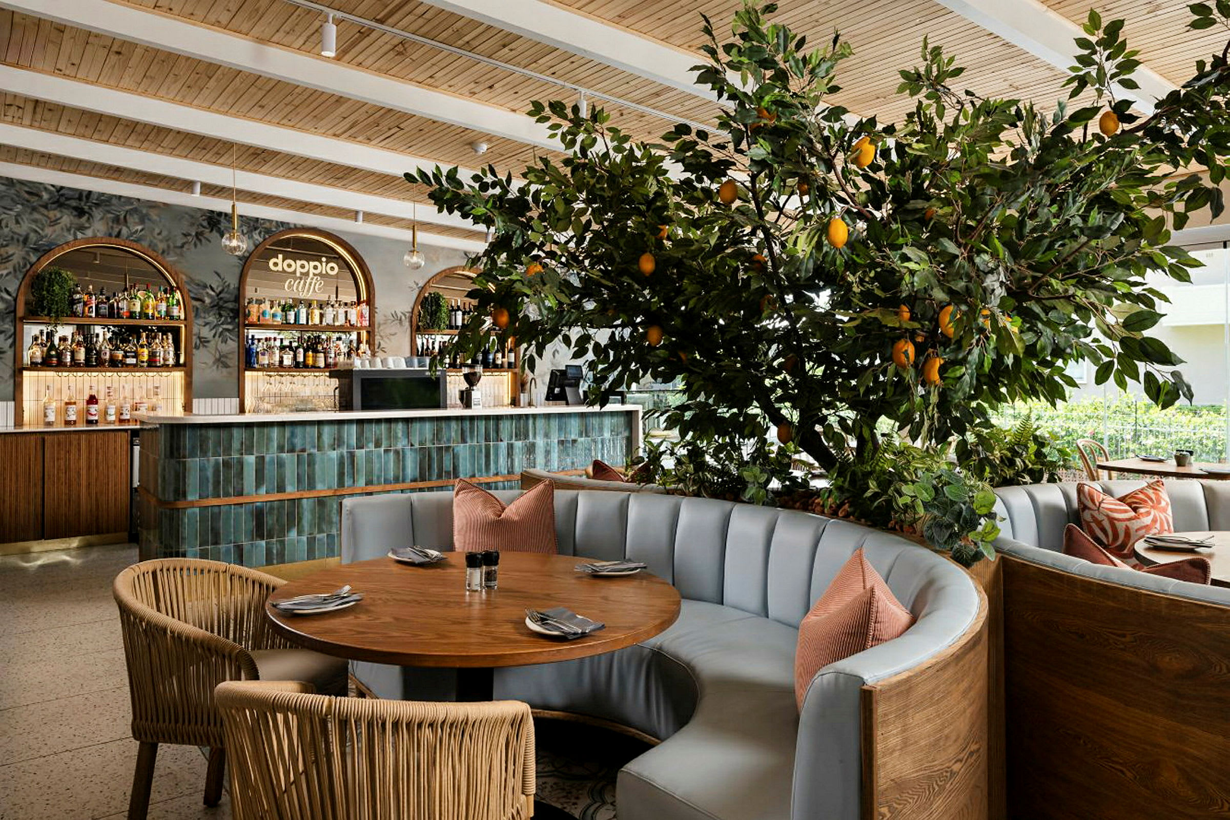 Interiør av en restaurant med et rundt bord, polstrede sofaer og en dekorativt plantet sitrusplante med frukt. I bakgrunnen sees en bardisk. (AI-generert tekst)