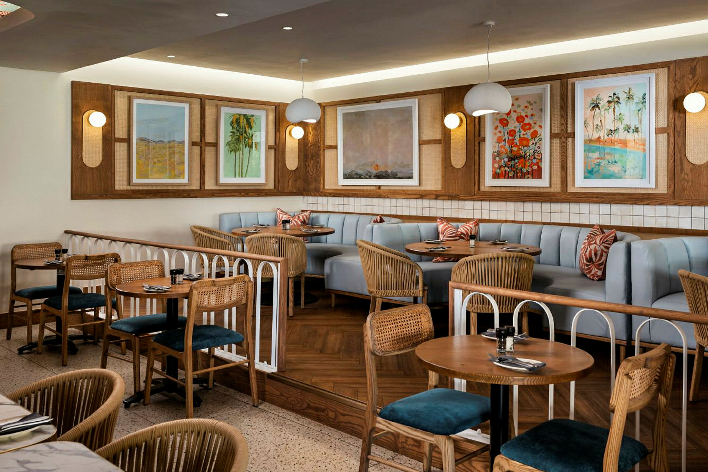 En restaurant med bord og stoler i tre og rotting samt en sittegruppe med lyseblå sofaer og dekorative puter. Veggene er utsmykket med malerier og lamper. (AI-generert tekst)