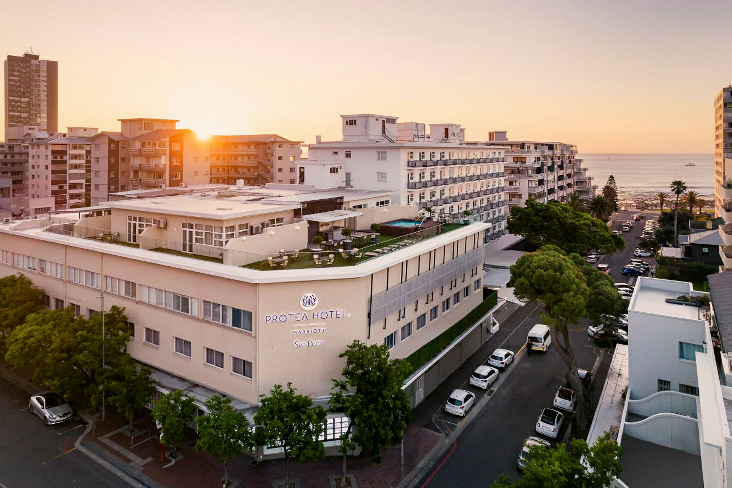 Hotellbygning ved sjøen omgitt av andre bygninger i solnedgang. (AI-generert tekst)
