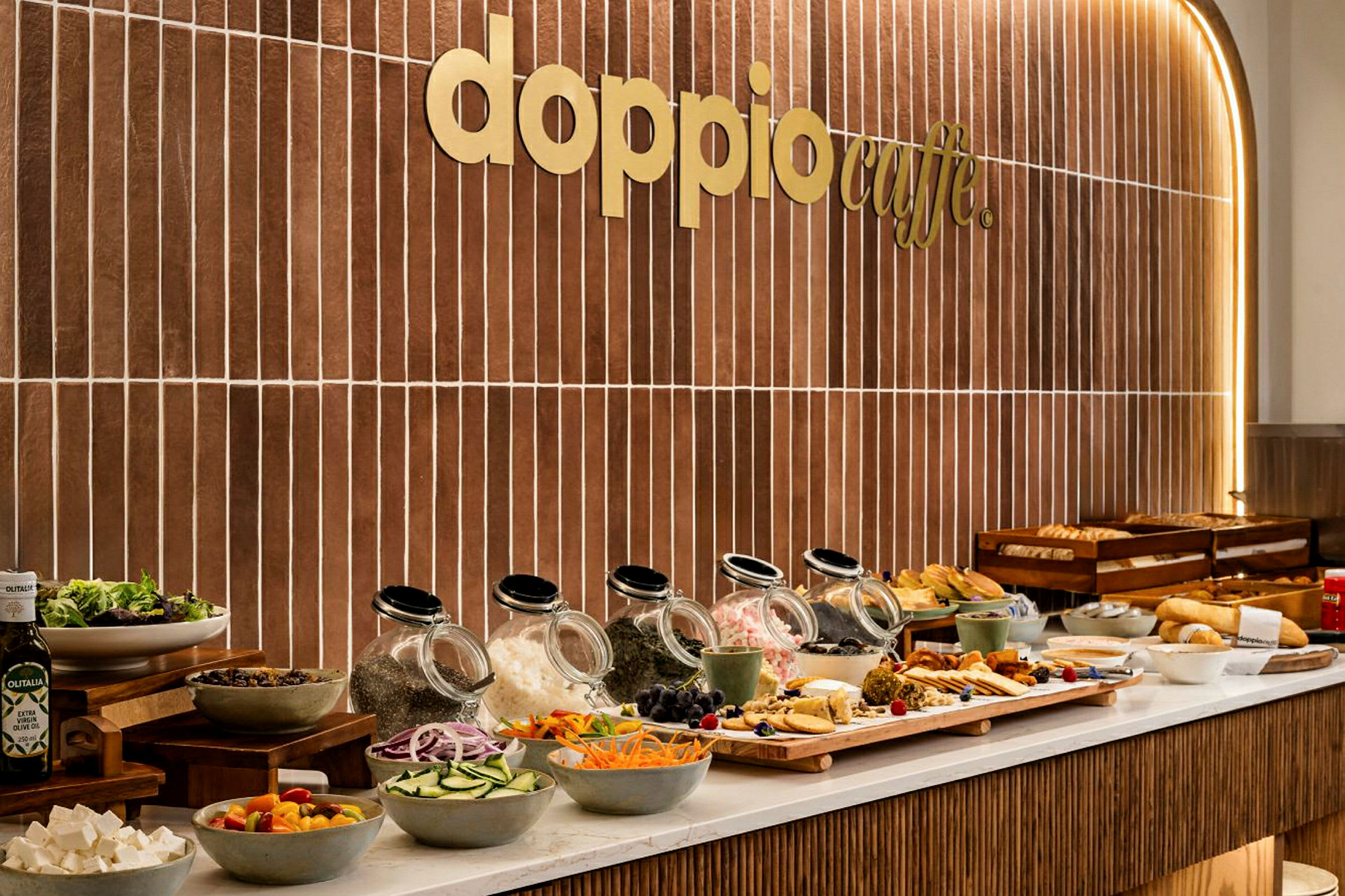 Bilde av en buffet med ulike matvarer og salatingredienser foran en vegg med teksten "Doppio Caffe". (AI-generert tekst)