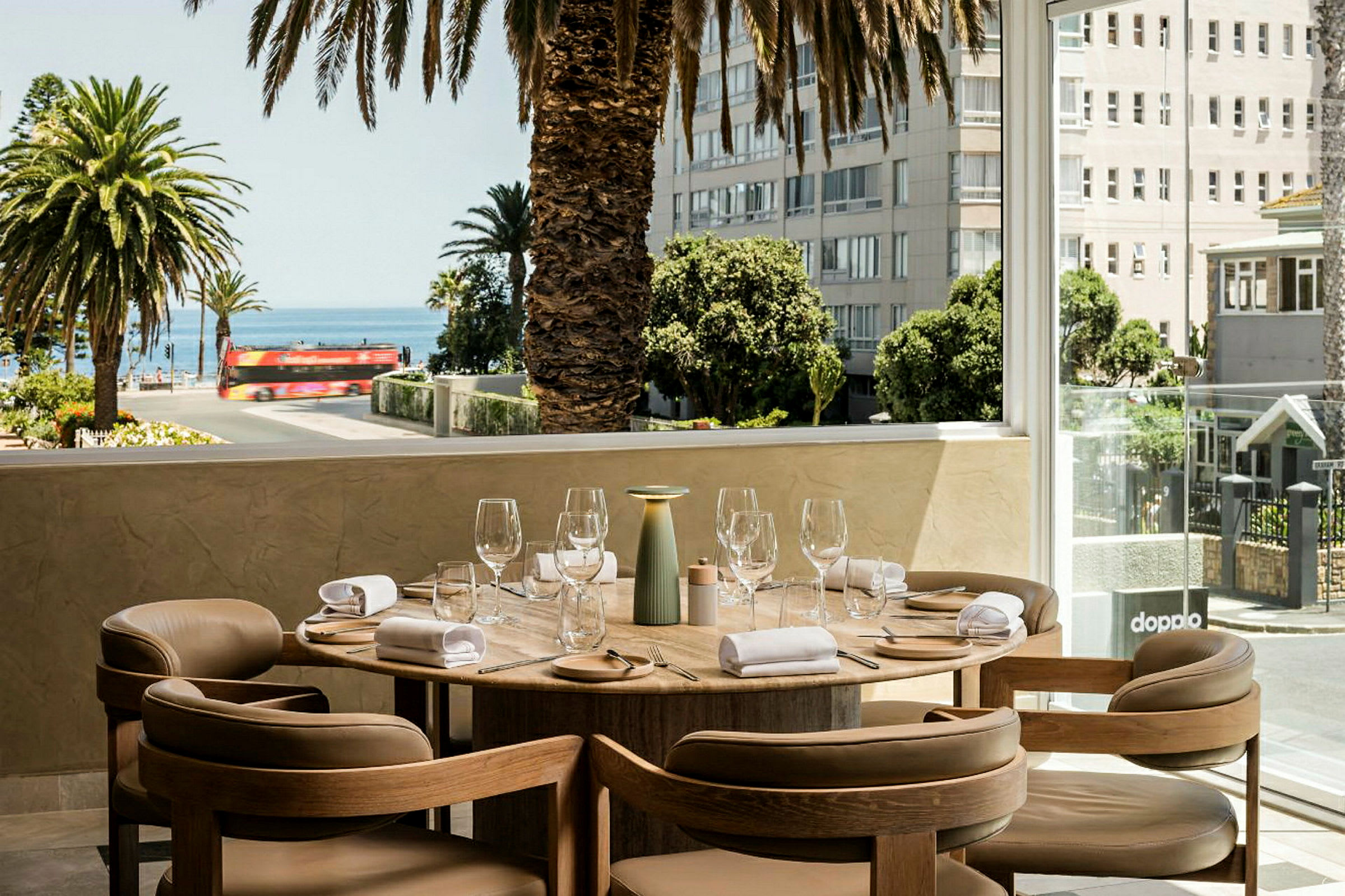 En dekket spiseplass på en terrasse med stoler rundt et rundt bord. Utsikten inkluderer palmer, vei, hav og bygninger i bakgrunnen. (AI-generert tekst)