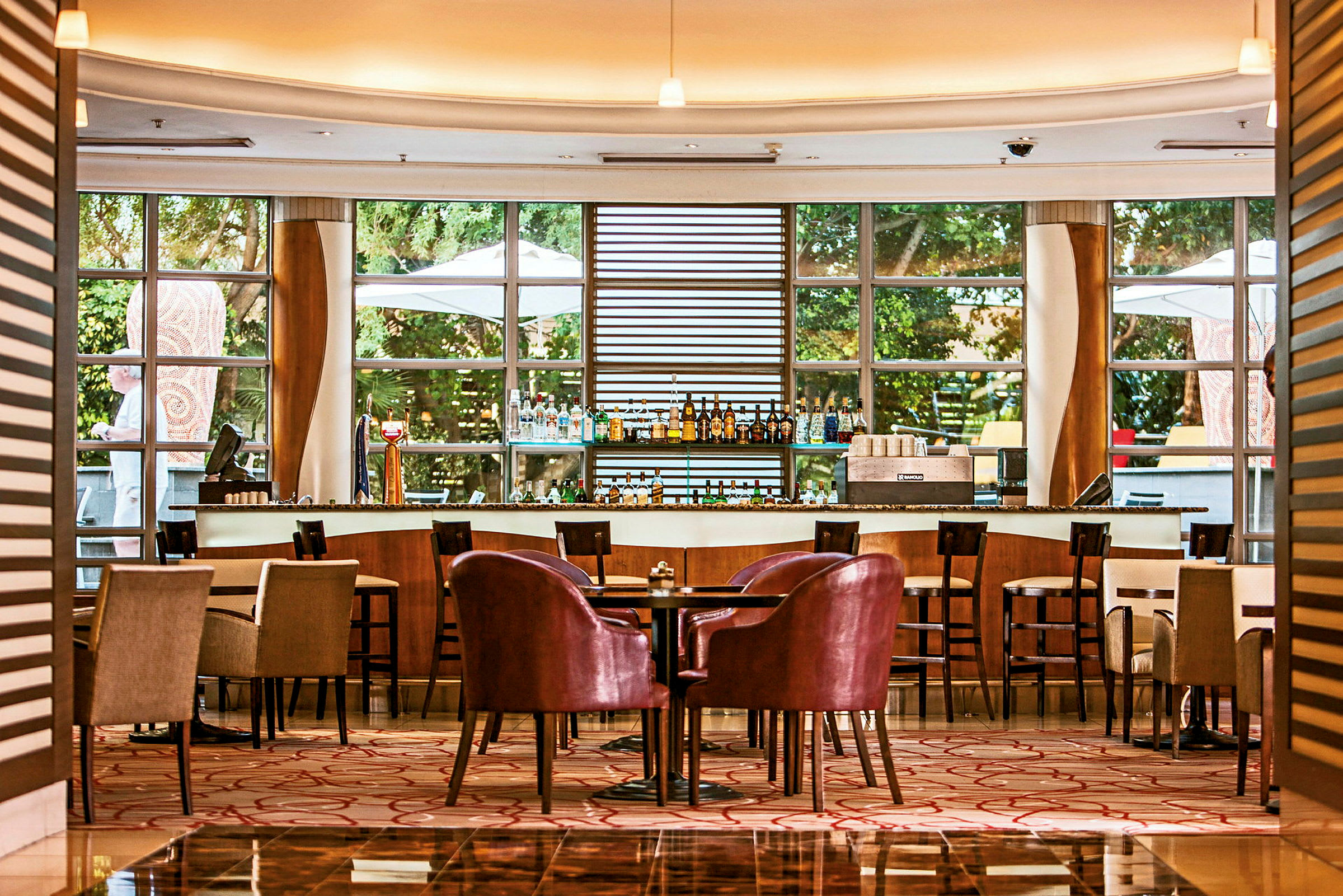 En elegant bar og sitteområde med bord og stoler på et hotell eller en restaurant. Store vinduer slipper inn mye lys utenfra. (AI-generert tekst)