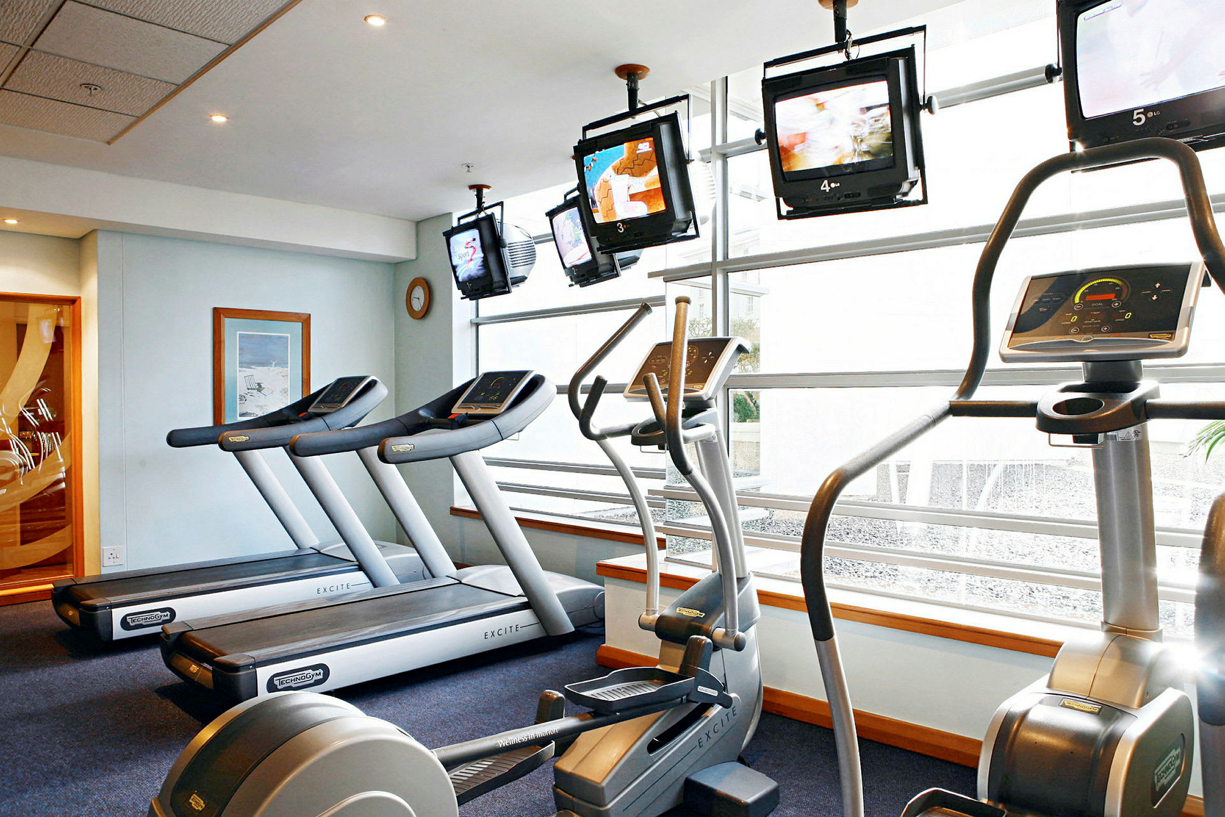 Bilde av et treningsstudio med treningsmaskiner som tredemøller og elliptiske maskiner, samt TV-skjermer nær vinduene. (AI-generert tekst)