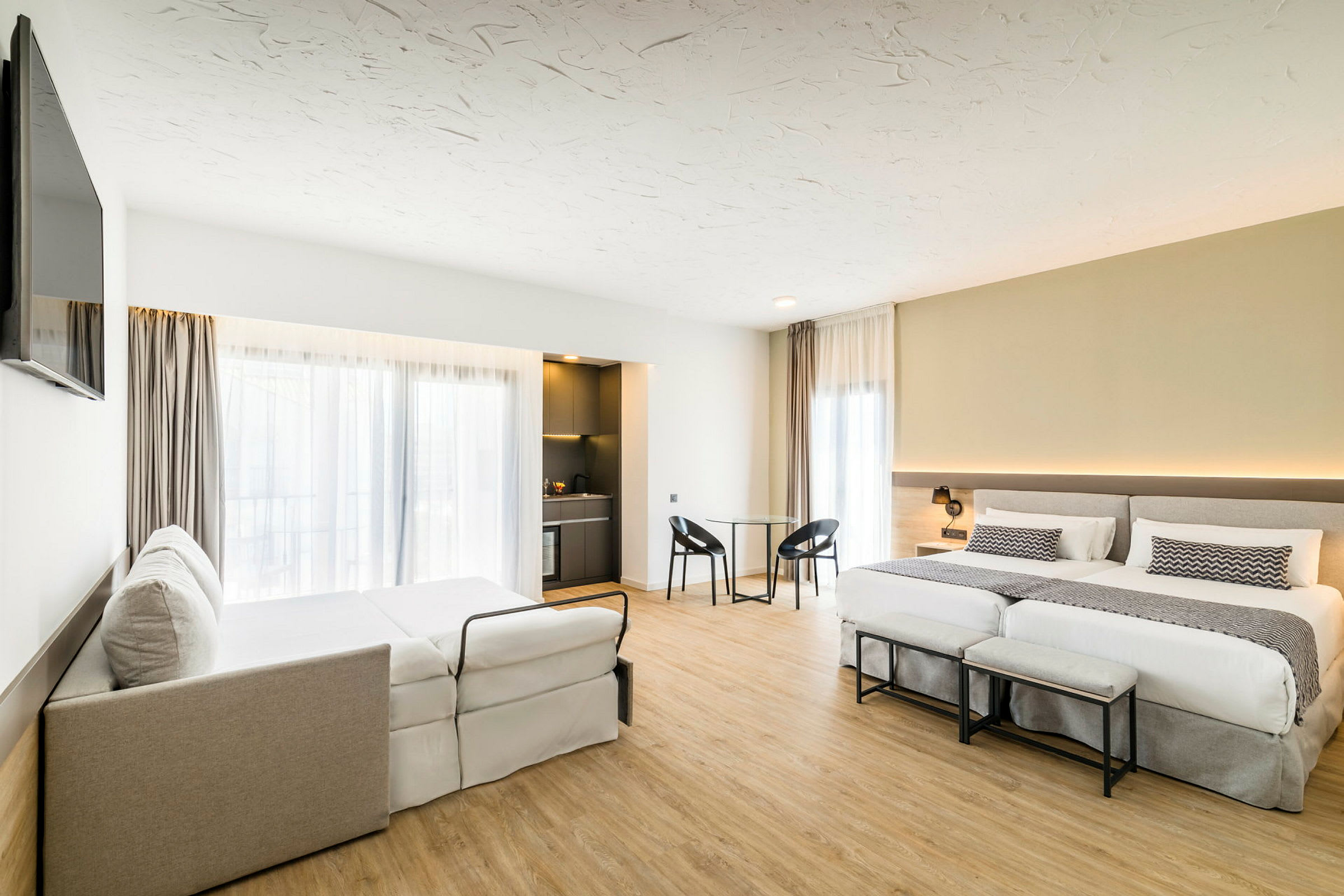 Hotellrom med to senger, en sovesofa, og et lite spisebord med stoler. Rommet har store vinduer med gardiner og en liten kjøkkenkrok. (AI-generert tekst)