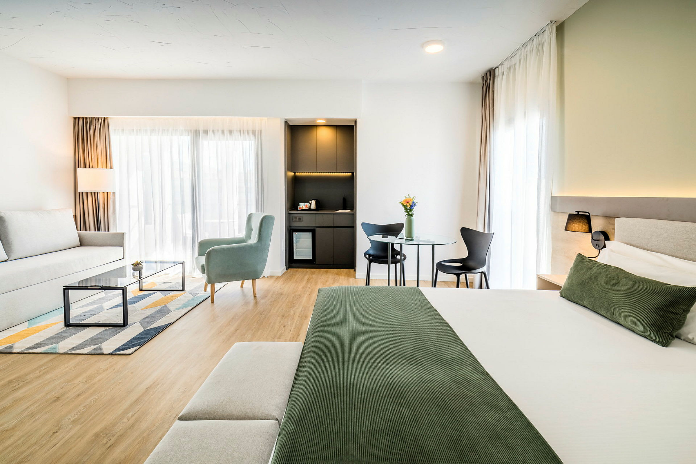 Et moderne innredet hotellrom med en seng, en sittegruppe, et bord med stoler og en liten kjøkkenkrok. (AI-generert tekst)