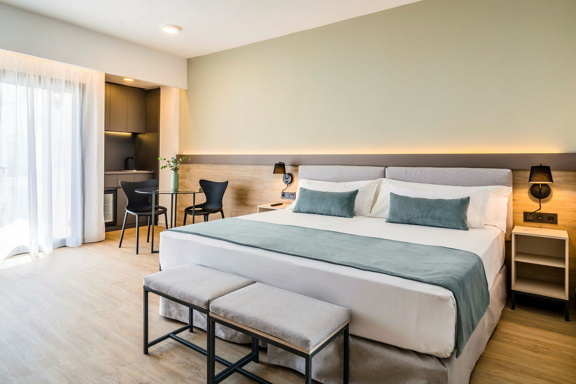 Et hotellrom med en stor dobbeltseng, to små benker ved fotenden, et sidebord og et bord med stoler nær et lite kjøkkenområde. (AI-generert tekst)