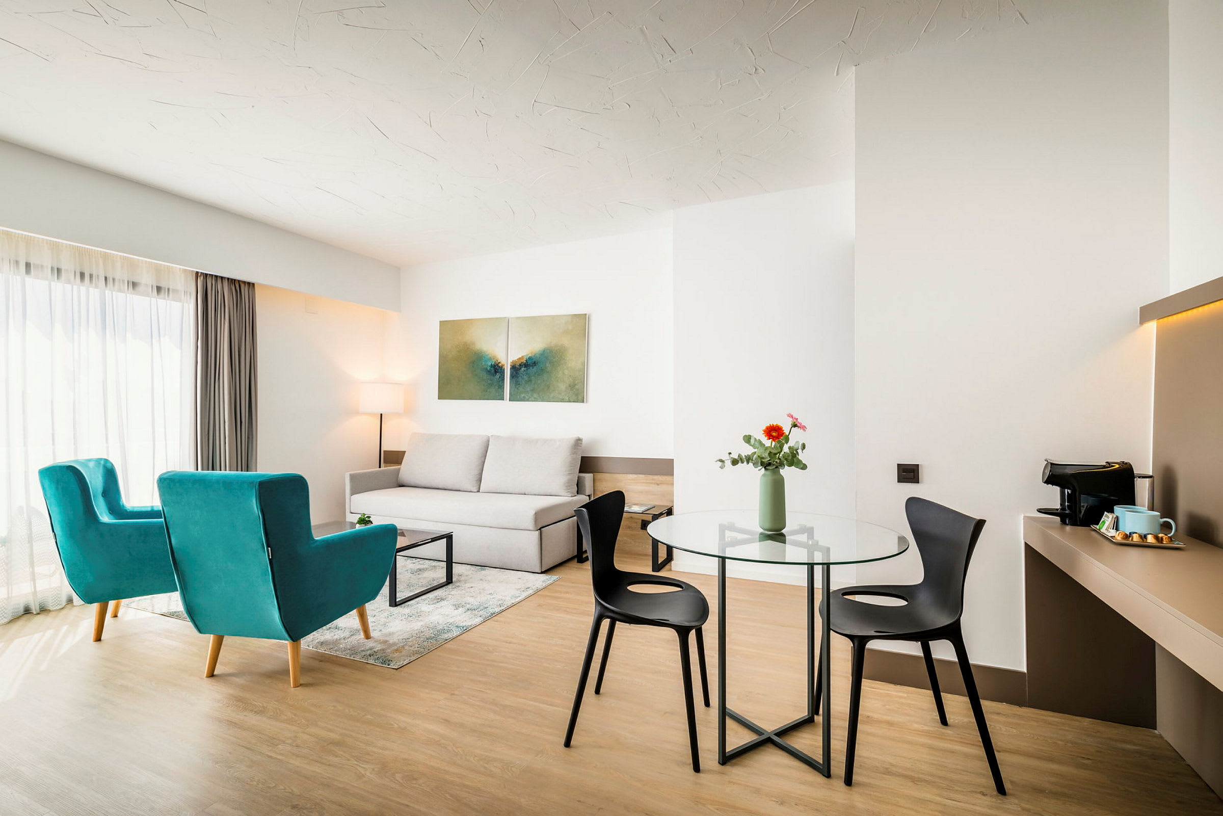 Et moderne rom med to blå lenestoler, en sofa, et glassbord med vase og to stoler. En kaffetrakter på en hylle er også synlig. (AI-generert tekst)
