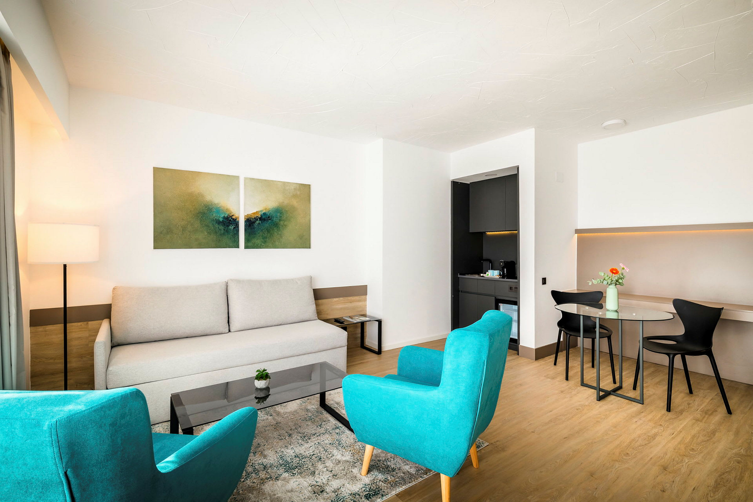 Et moderne hotellrom med sittegruppe, to turkise lenestoler, en lysegrå sofa og et glassbord. Et lite kjøkkenområde vises til høyre. (AI-generert tekst)