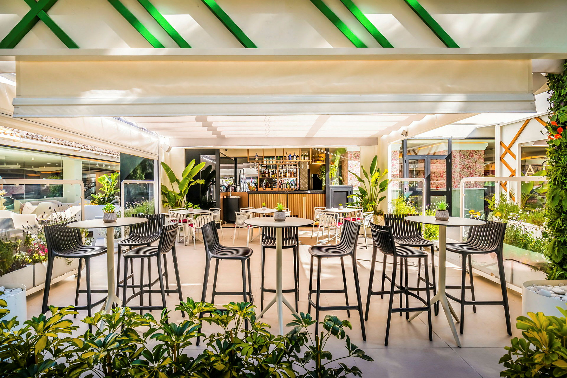 En moderne restaurant med uteservering, omgitt av planter og med høye barstoler rundt små bord. (AI-generert tekst)
