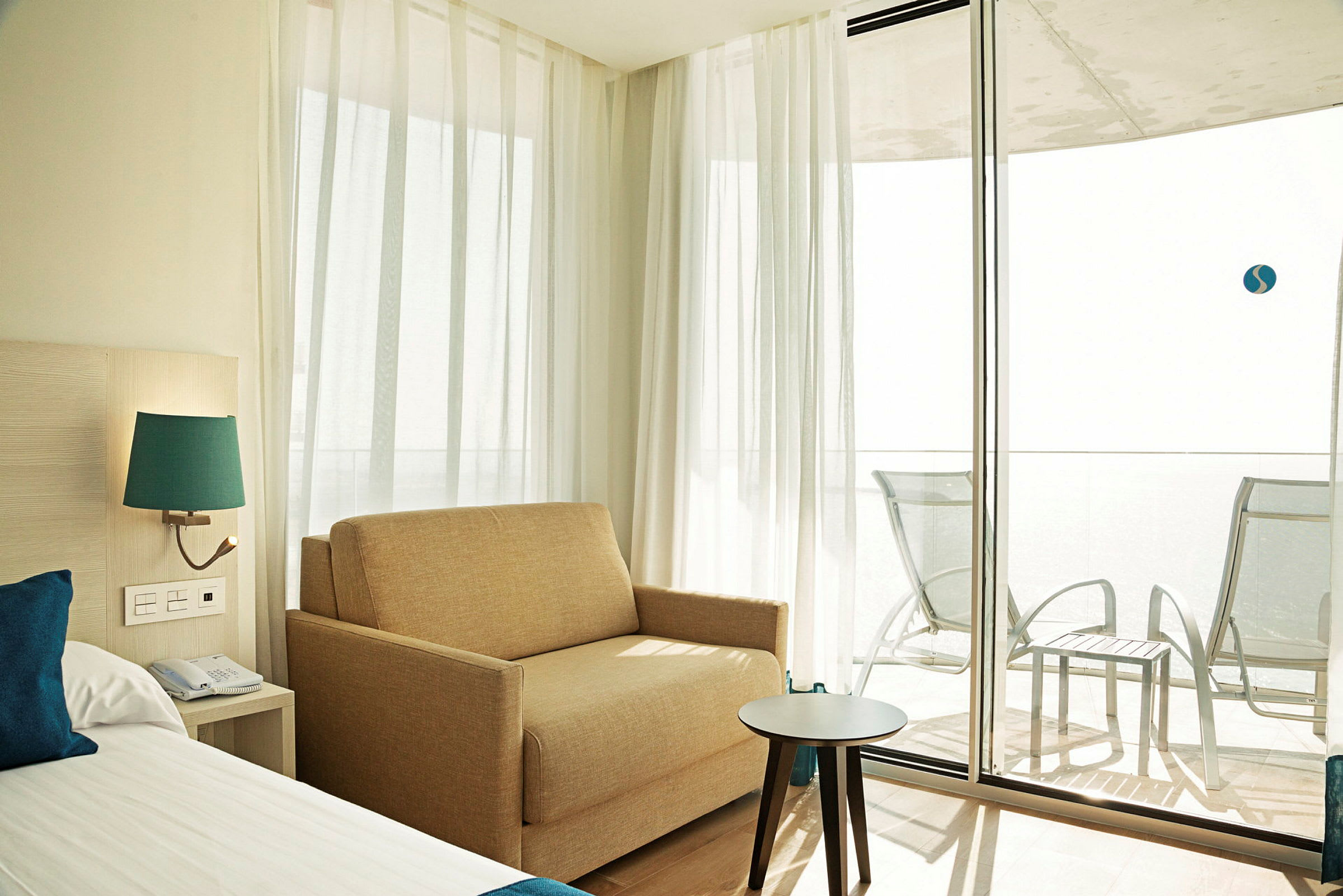 Et hotellrom med en seng, en sofa og et bord nær glassdører som fører til en møblert balkong. (AI-generert tekst)