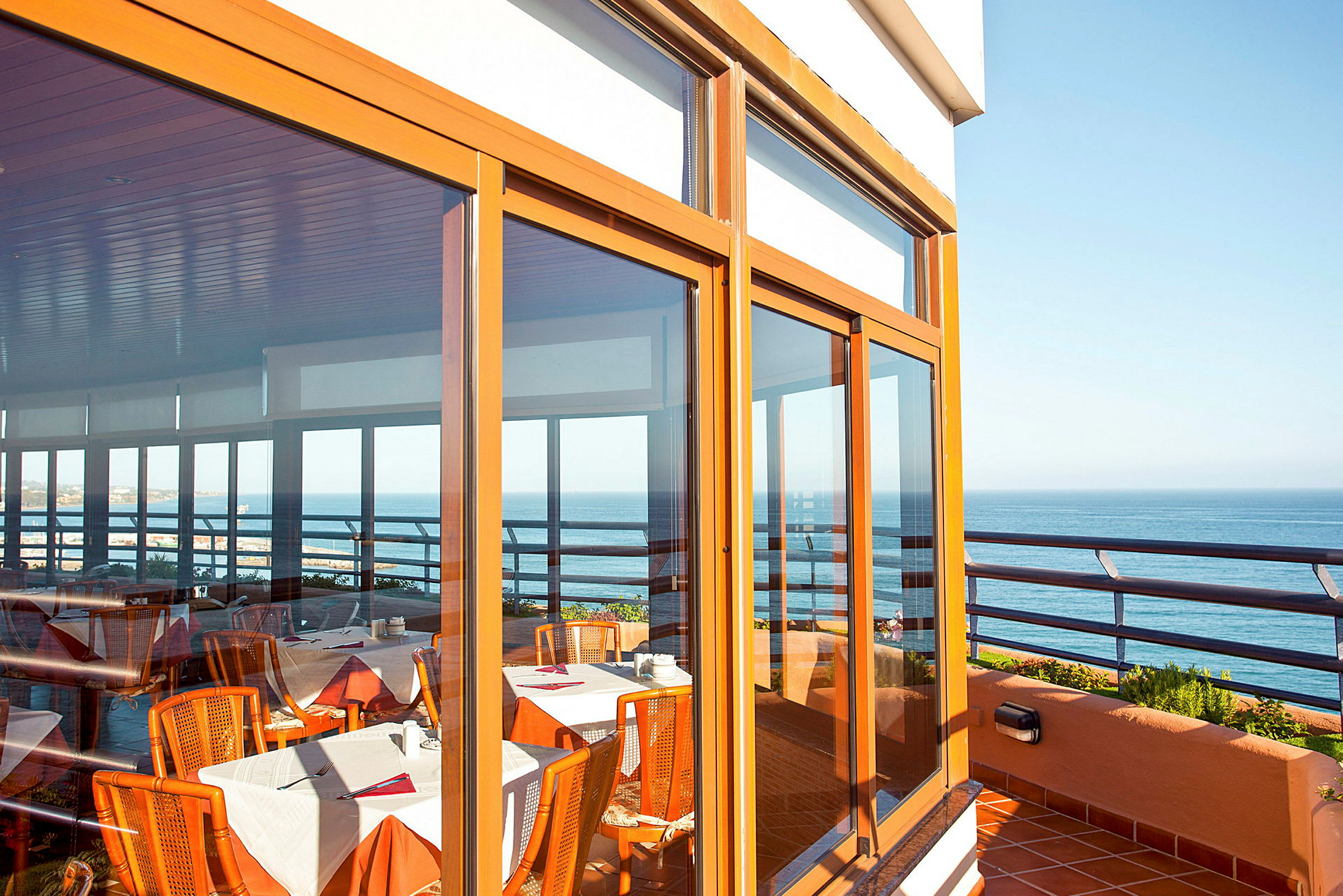 Utsikt over en terrasse med glasspartier vendt mot havet. Innendørs er det bord med duker og stoler. (AI-generert tekst)