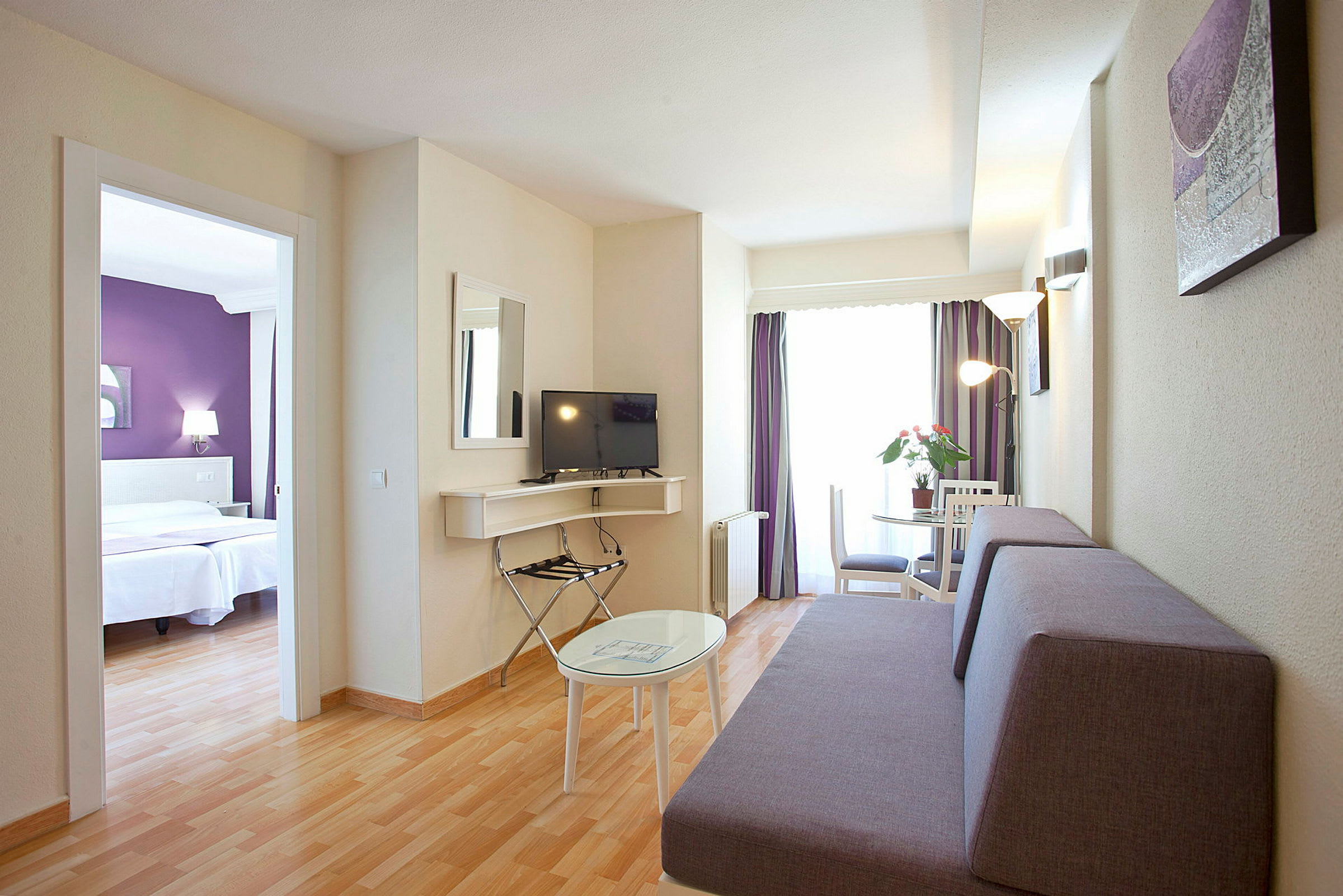 Hotellrom med stueområde inkludert sofa, TV og bord, samt separat soverom synlig gjennom en døråpning. (AI-generert tekst)
