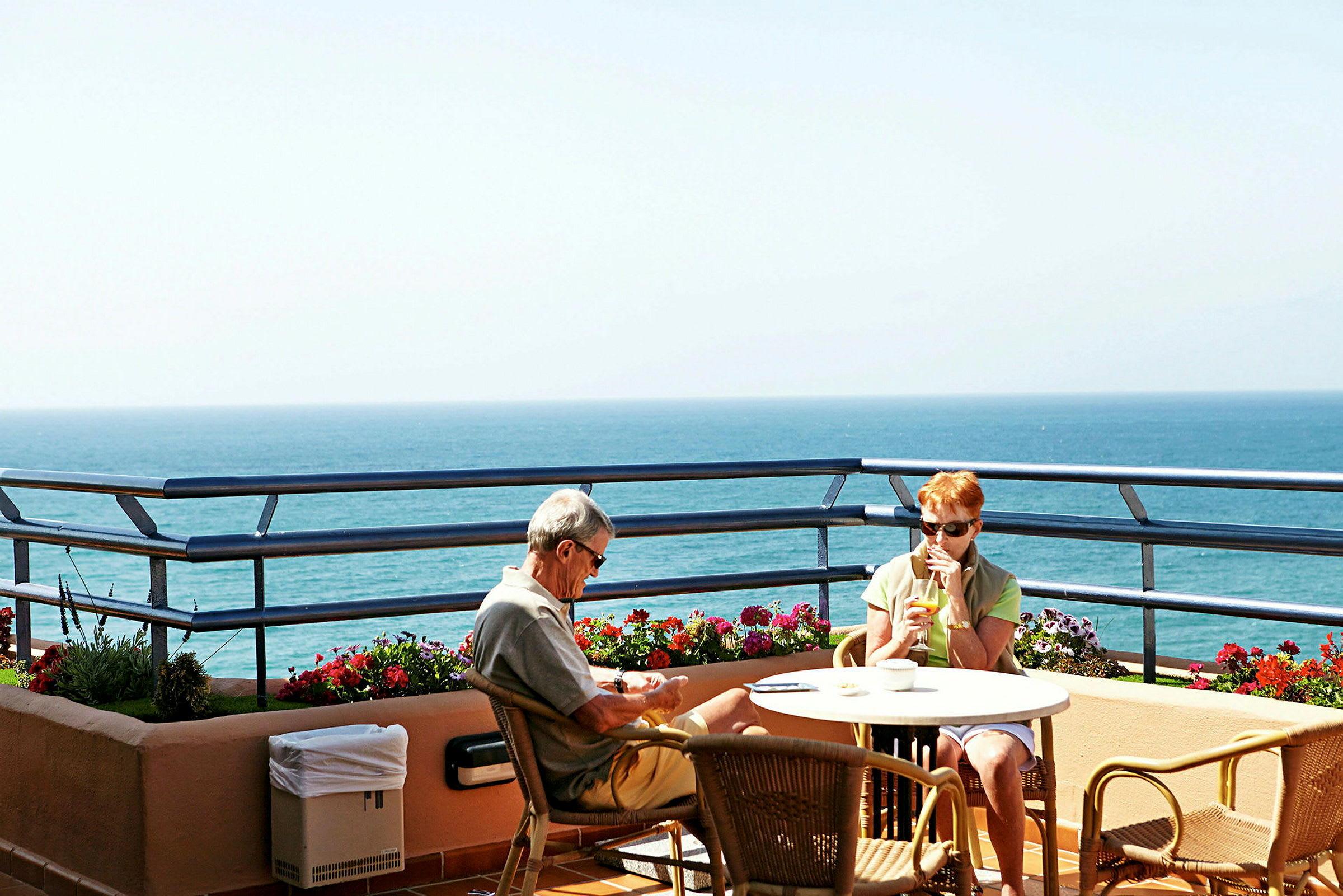 To personer sitter på en terrasse med utsikt over havet. (AI-generert tekst)