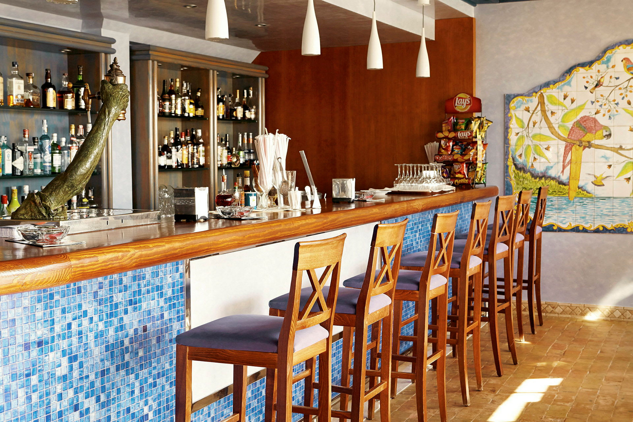 En bar med høye trestoler, en mosaikkfront i blått, et imponerende utvalg av spritflasker og en veggdekorasjon med en papegøye. (AI-generert tekst)