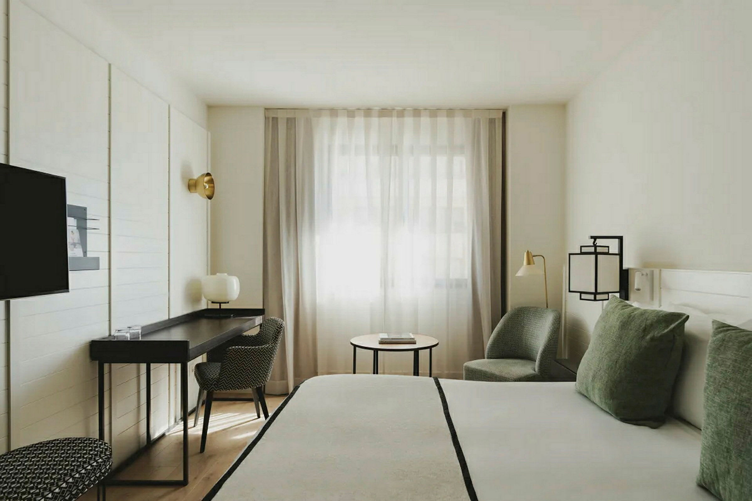 Et lyst hotellrom med en stor seng, skrivebord, stol og lenestol. Vinduet har lette gardiner. (AI-generert tekst)