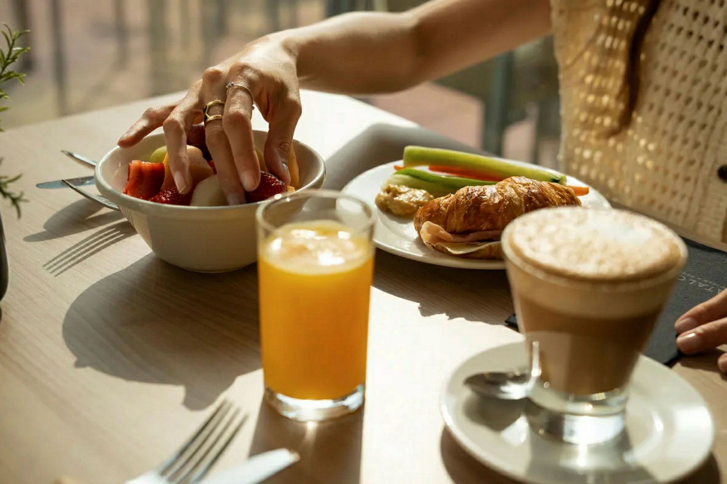 Person tar frukt fra en skål på et bord med croissant, grønnsaker, juice og kaffe. (AI-generert tekst)