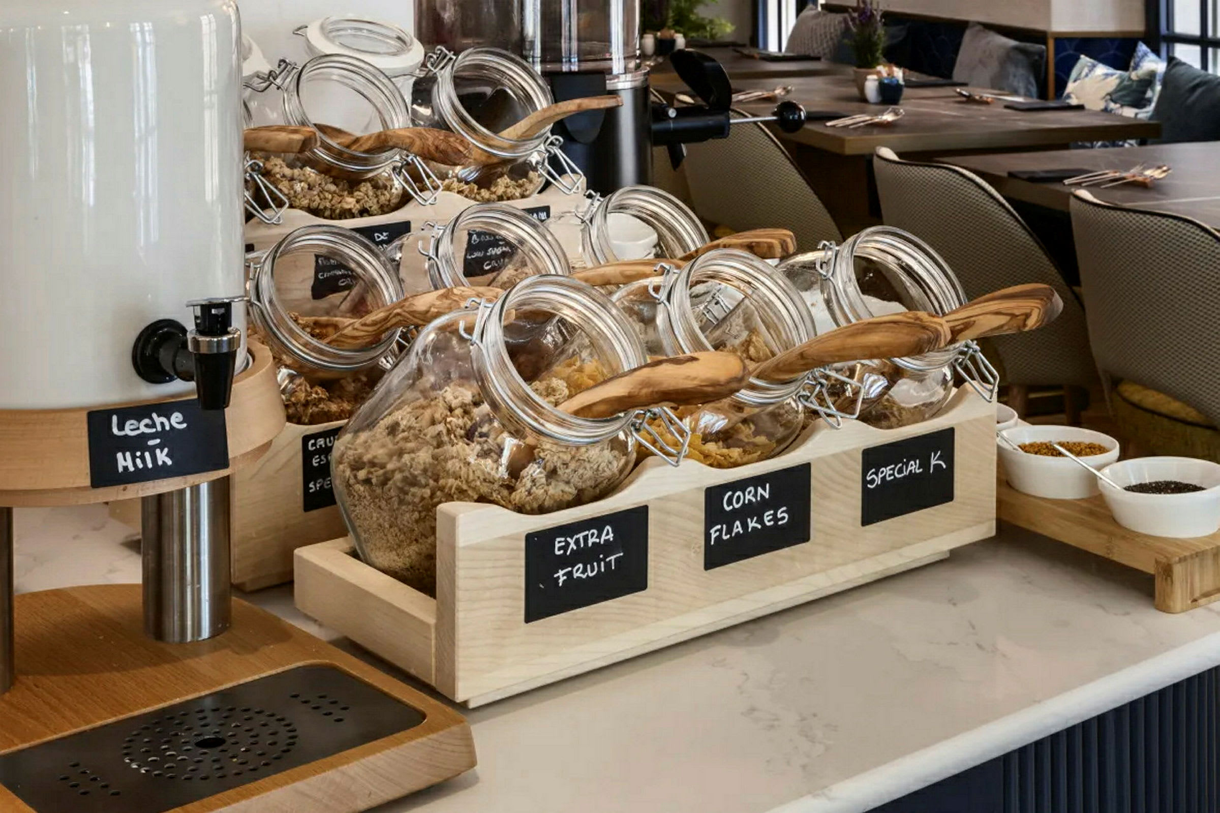 Glasskrukker fylt med forskjellige typer frokostblandinger og müsliblandinger på et hotelldisk med melkebeholder og skåler med pålegg. (AI-generert tekst)