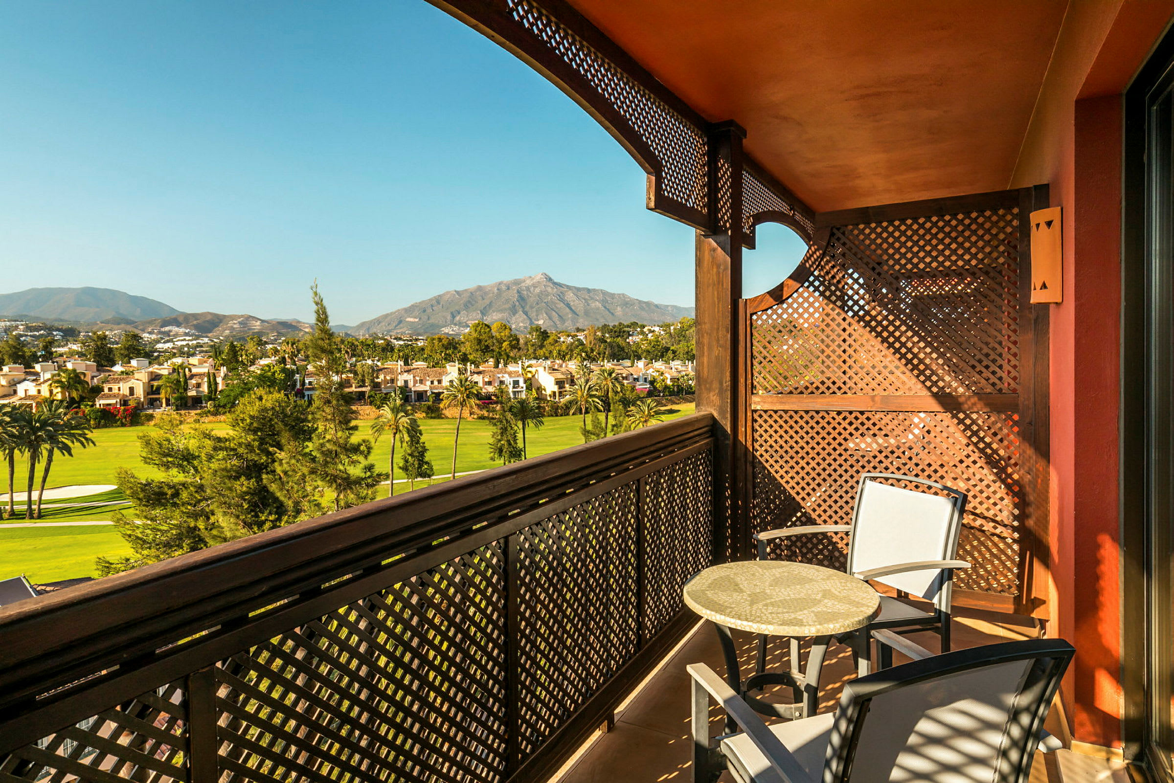 Balkong med to stoler og et bord, med utsikt over grønne trær, hus og fjell. (AI-generert tekst)