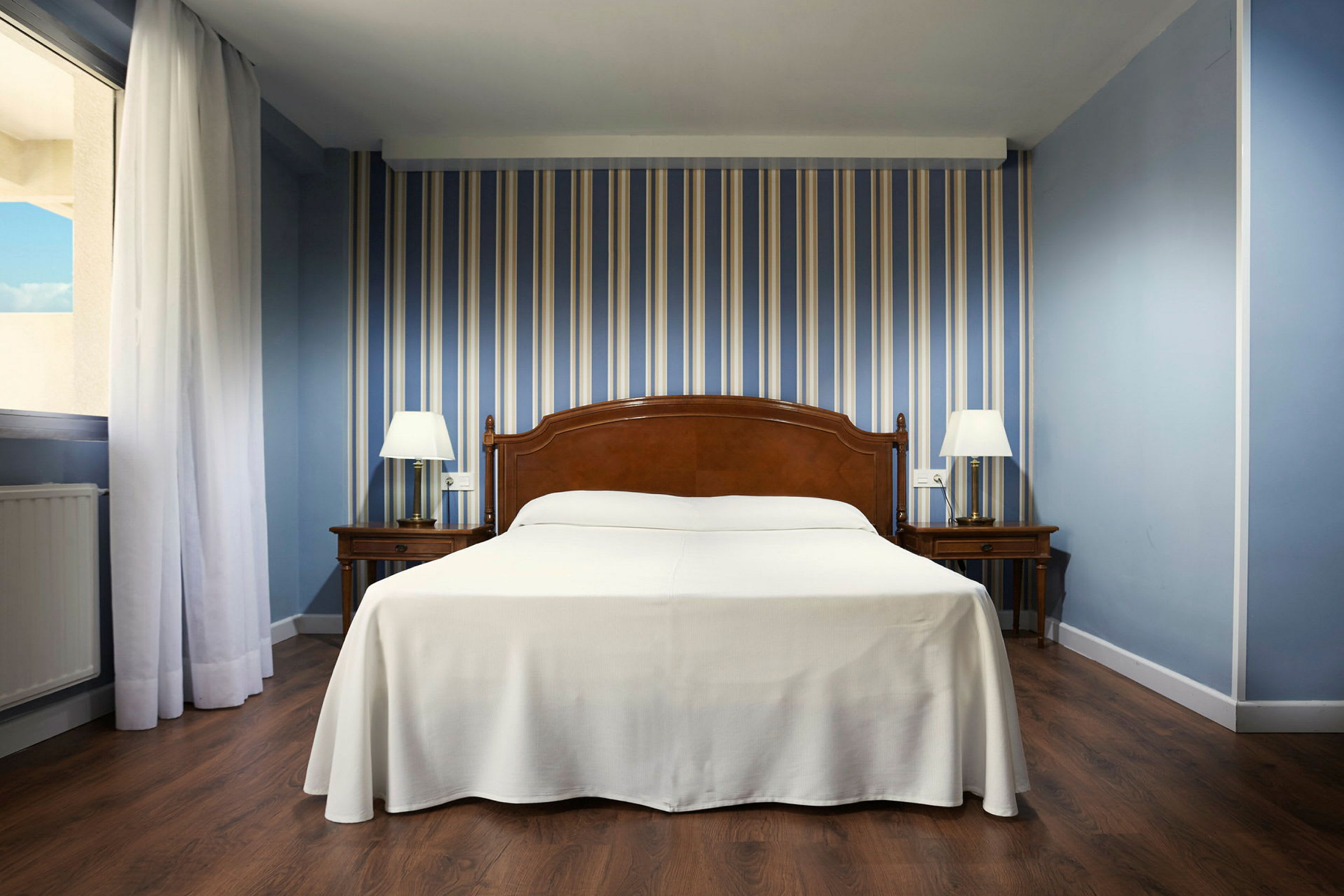 Hotellrom med seng i midten, to nattbord med lamper og stripete blå tapet. (AI-generert tekst)