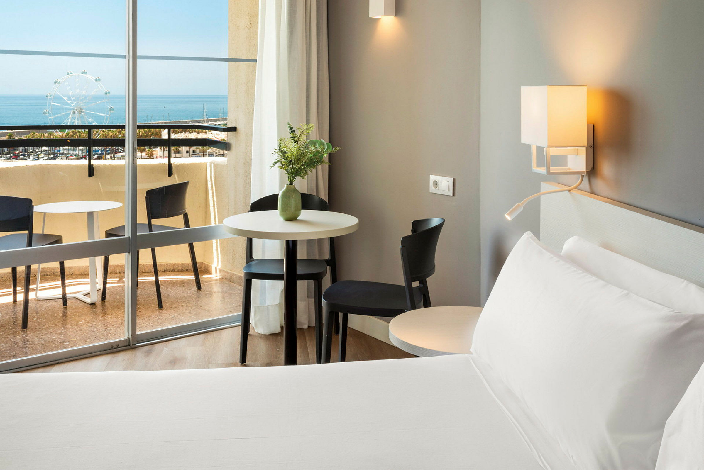 Et hotellrom med en seng, bord og stoler nær en balkong med utsikt over havet. (AI-generert tekst)