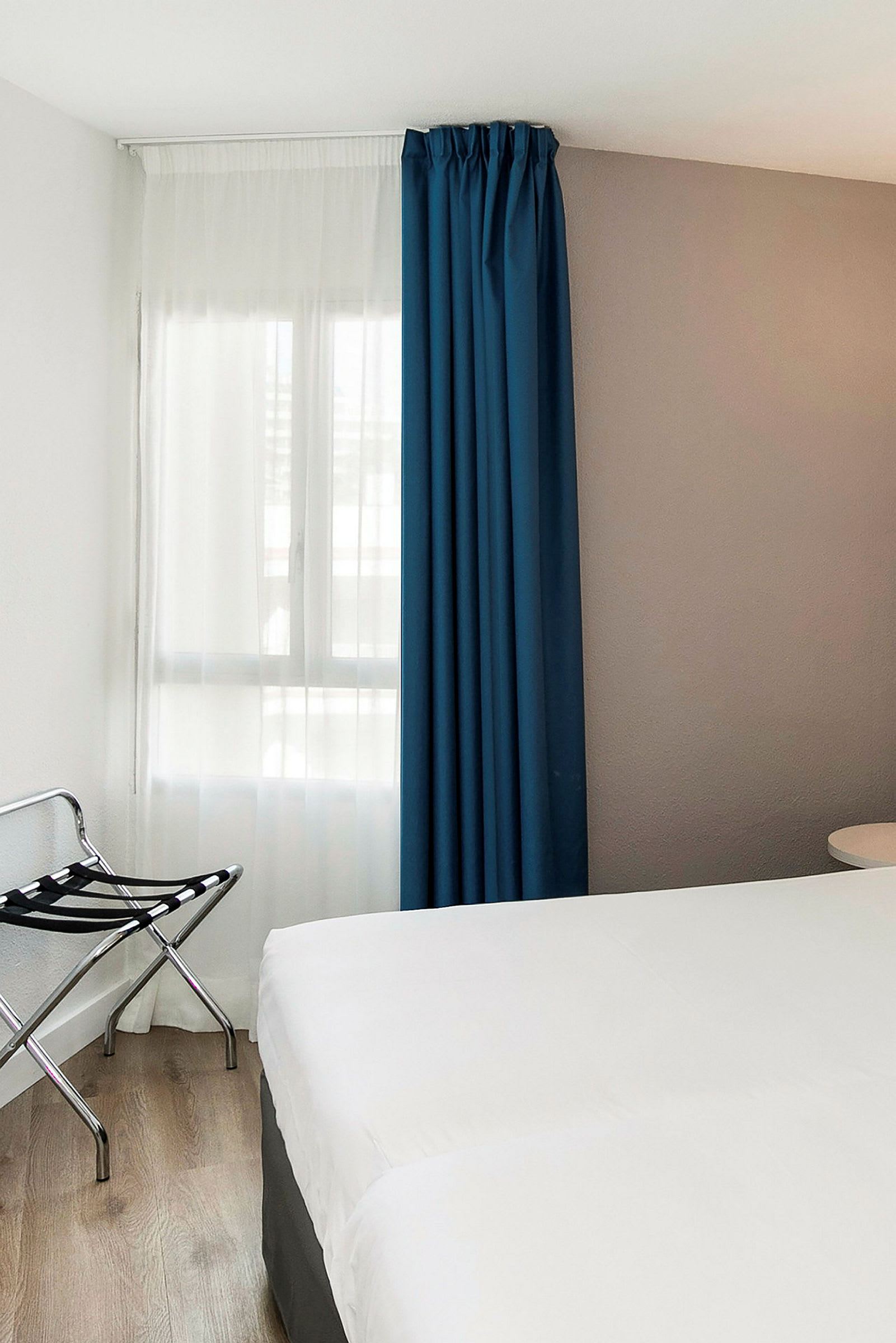 Et hotellrom med en hvit sengeteppe, en blå gardin og en bagasjeholder ved siden. (AI-generert tekst)